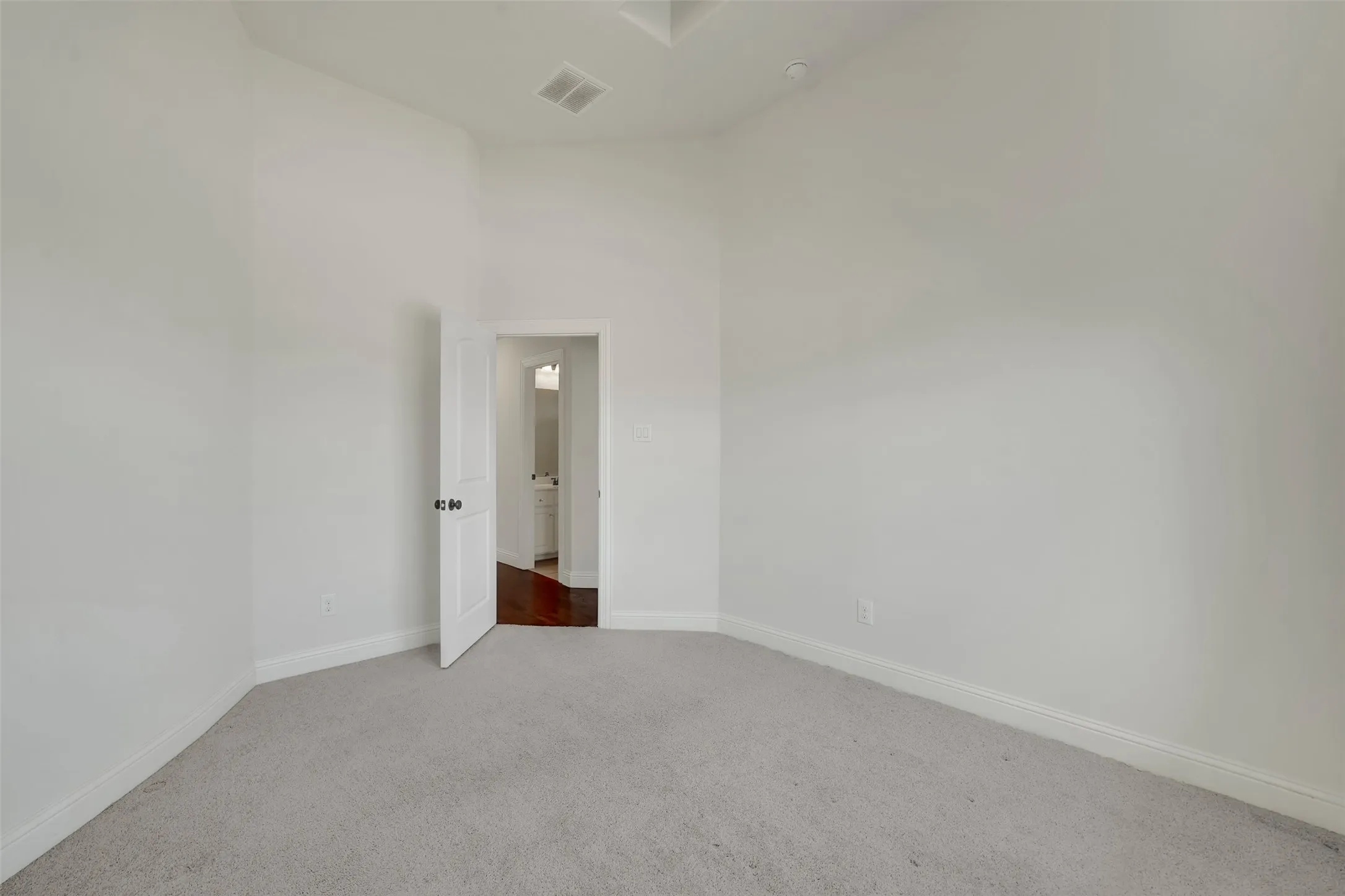 Vaulted ceiling, light neutral wall color, plush carpet flooring, white interior door, and dark wood-finish flooring visible through doorway