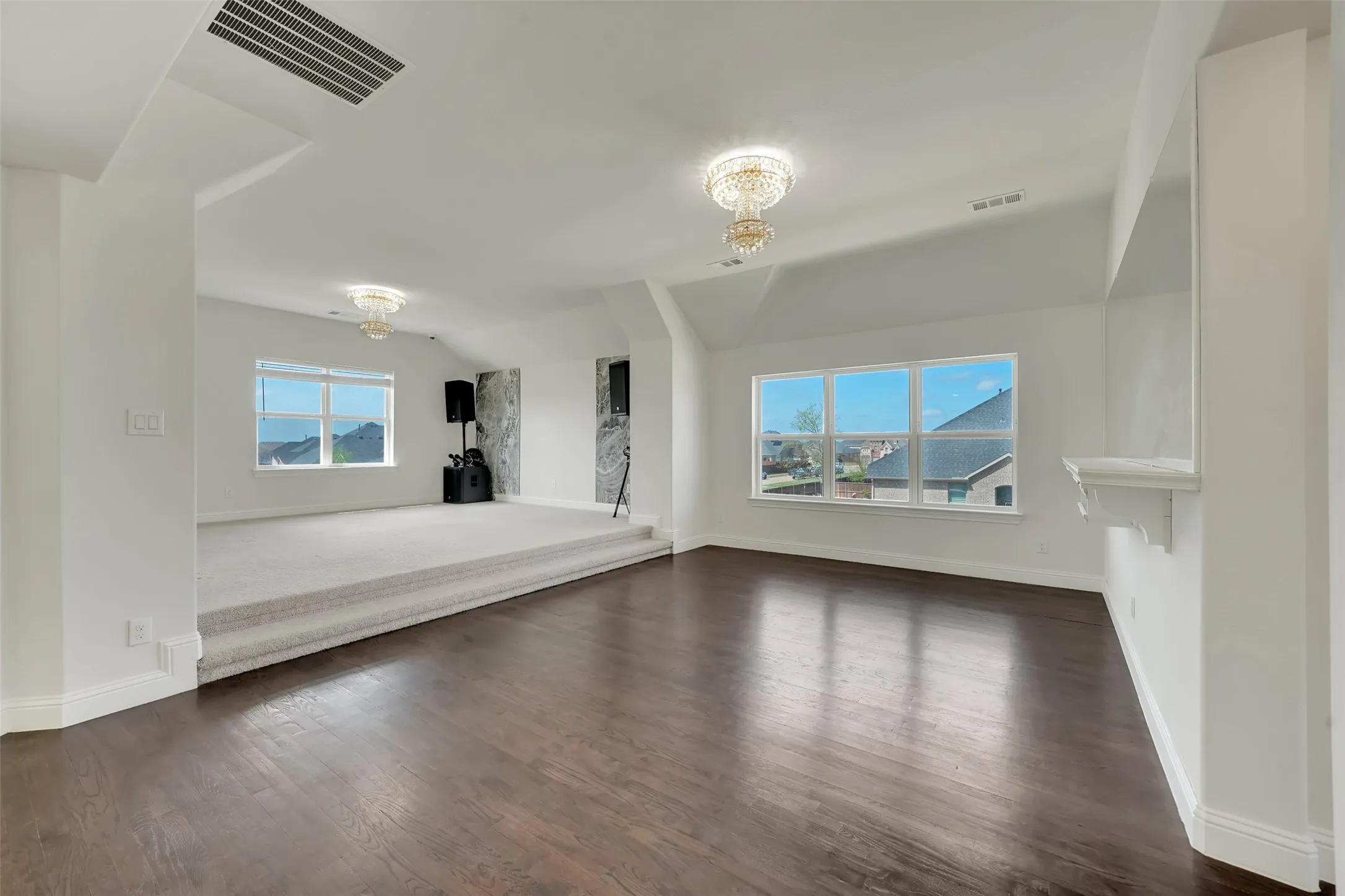 Spacious room featuring wood-finish flooring, a raised carpeted area, multiple windows, and two ornate chandeliers