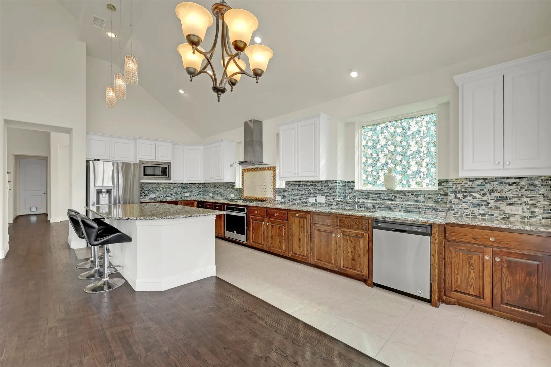 Vaulted ceilings, a kitchen island with granite countertops, stainless steel appliances, white upper cabinetry, and wood-finish lower cabinetry