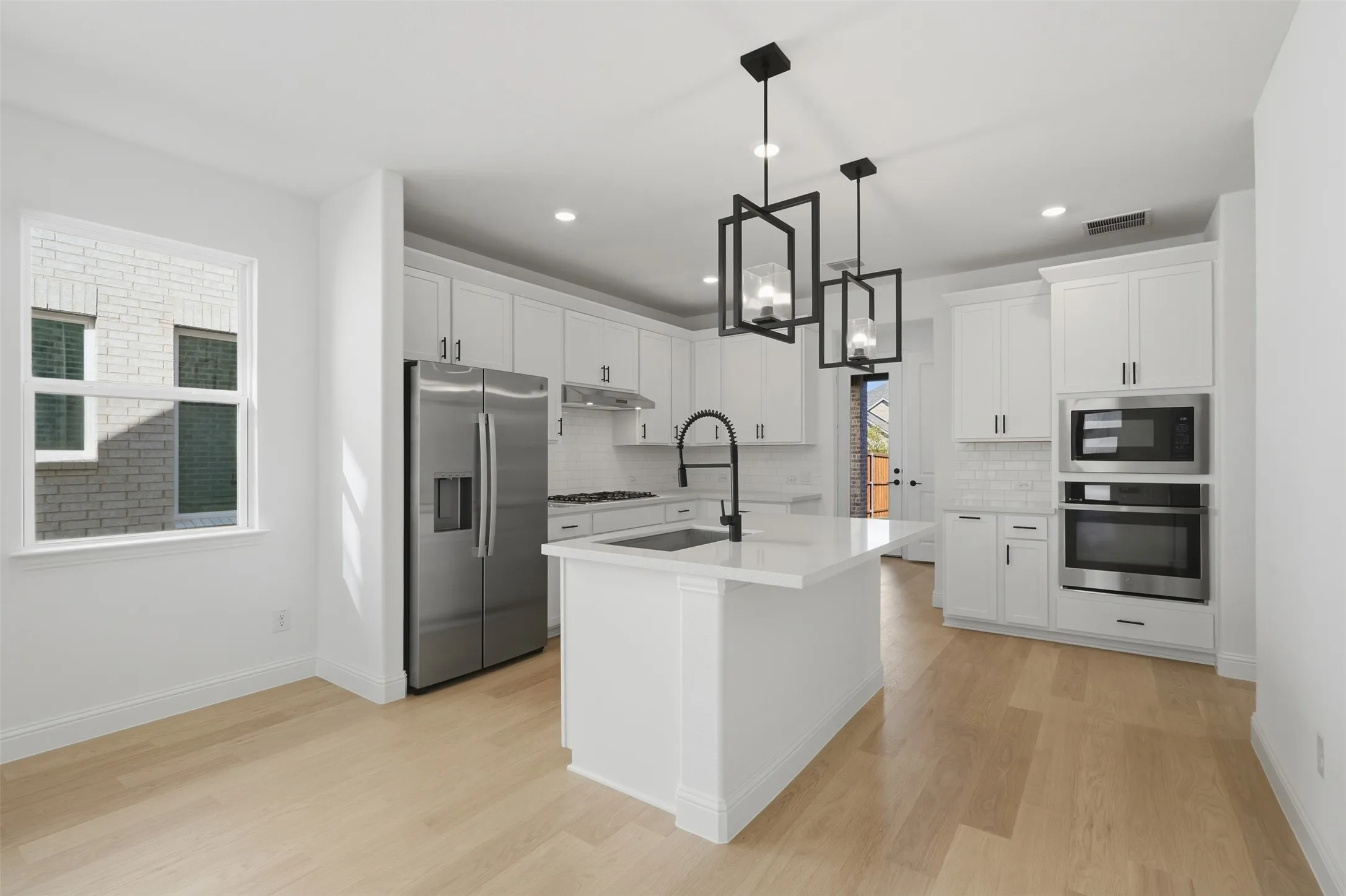 Open-concept kitchen featuring an island with a sink, stainless steel appliances, white cabinetry, wood-finish flooring, and two contemporary pendant lights
