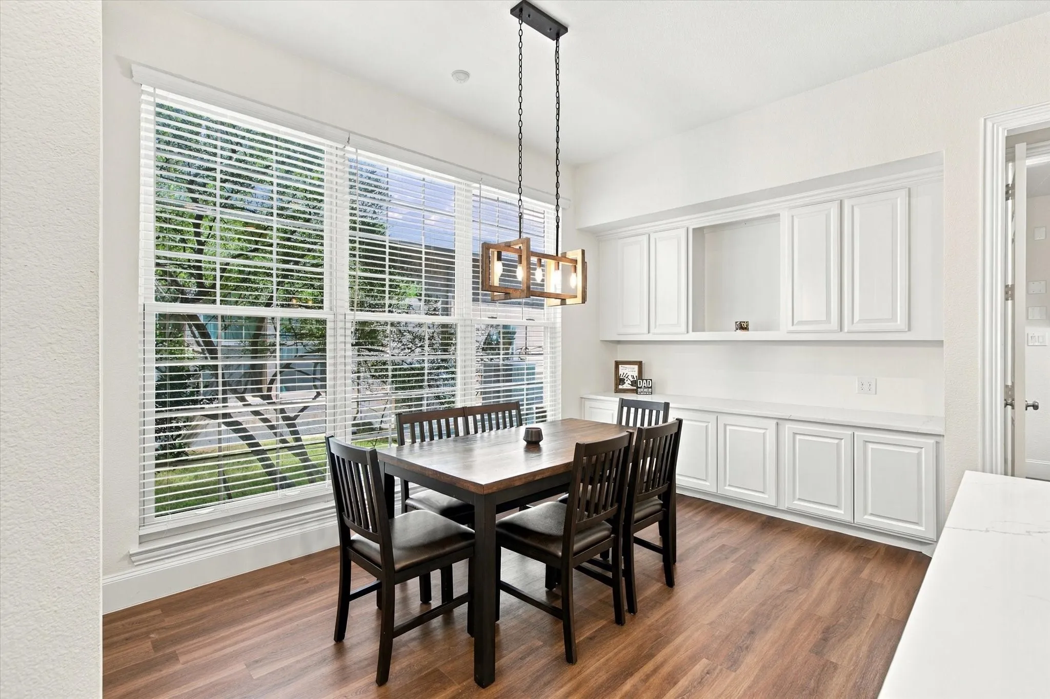 Wood-finish flooring, large multi-pane window with blinds, built-in cabinetry with white shaker-style doors, and a decorative chandelier