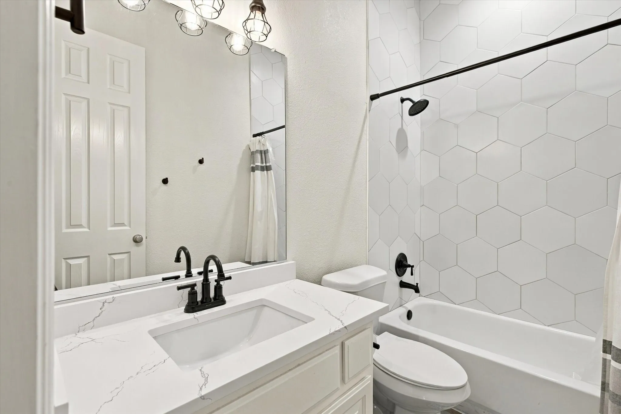 White hexagonal tile shower surround, matte black shower fixtures, white vanity with integrated rectangular sink, quartz-finish countertop, and matte black faucet