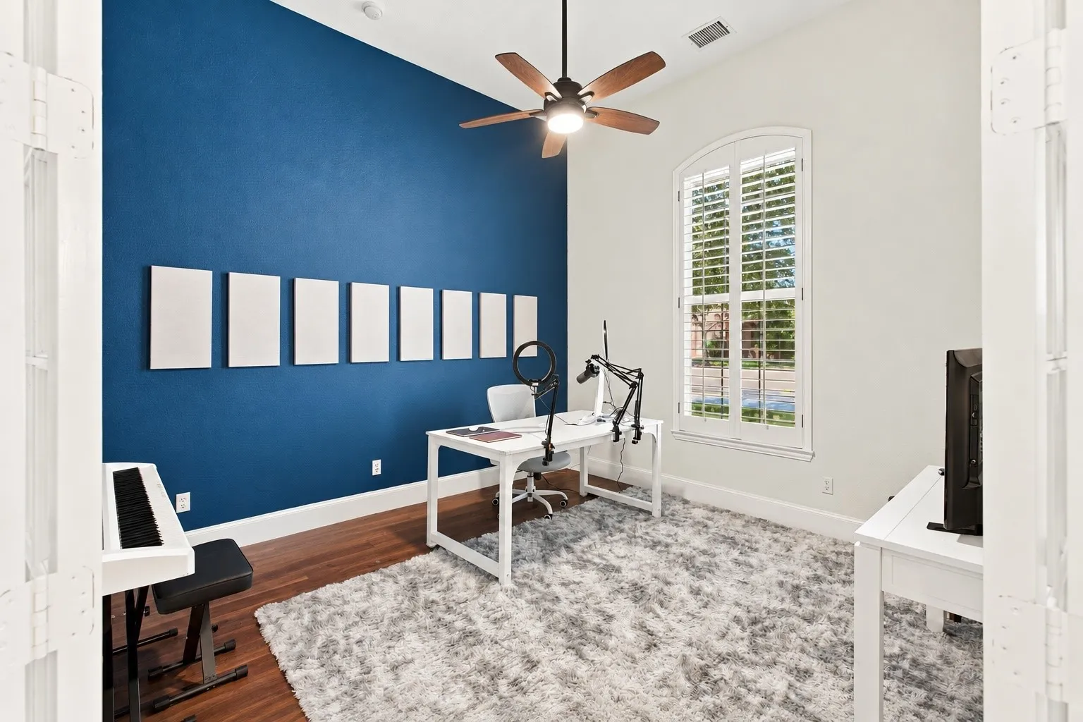 Vaulted ceiling with wood-finish ceiling fan, arched window with plantation shutters, wood-finish flooring, and a distinct accent wall