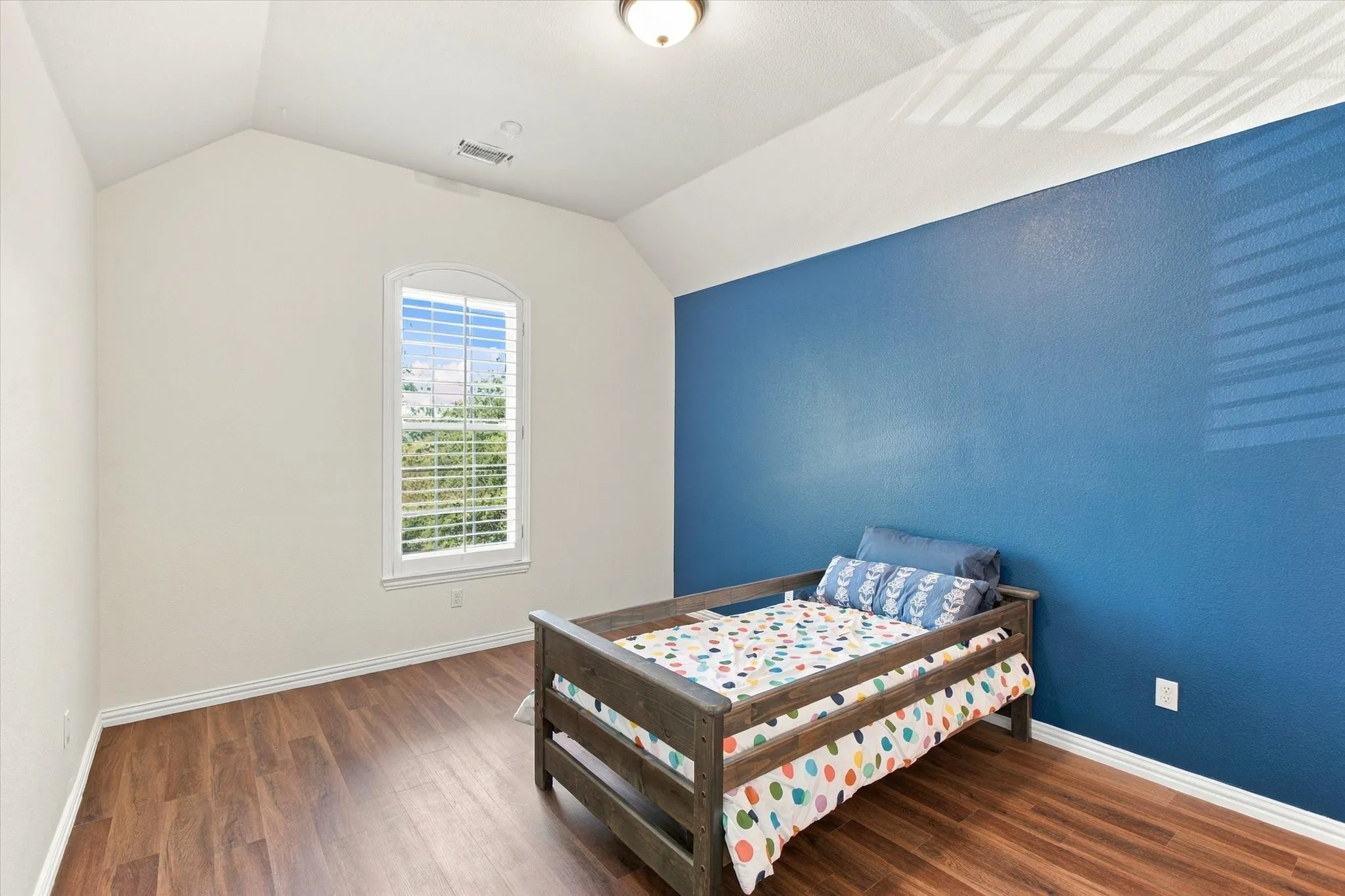 Vaulted ceiling, arched window with plantation shutters, wood-finish flooring, and a flush mount ceiling light