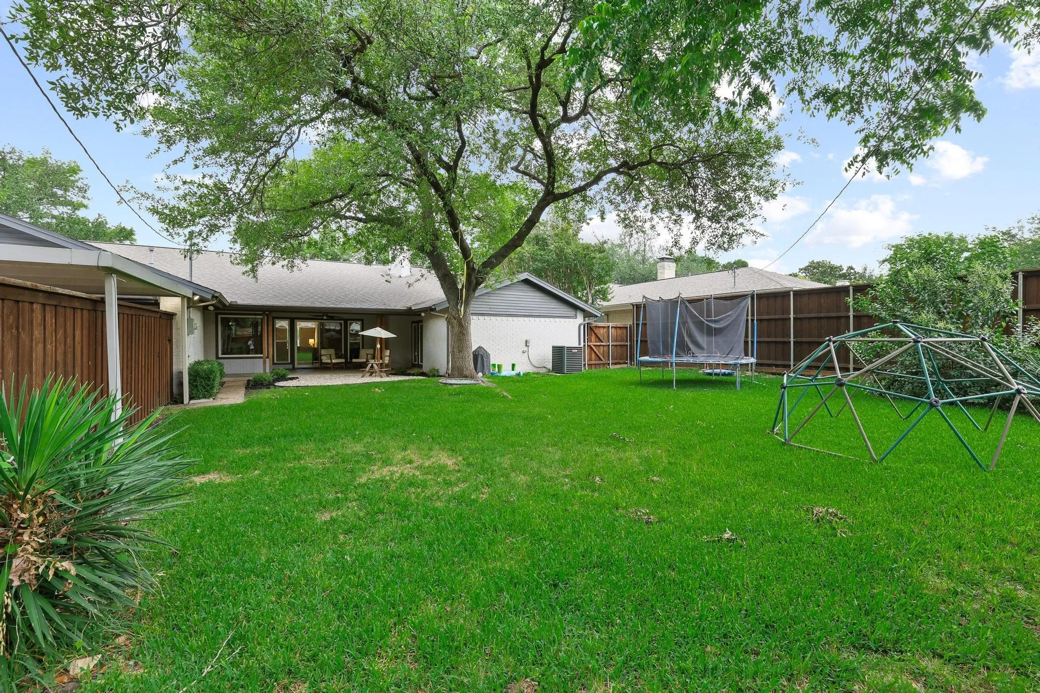 Expansive backyard featuring a manicured lawn, mature shade tree, privacy fencing, and an outdoor patio