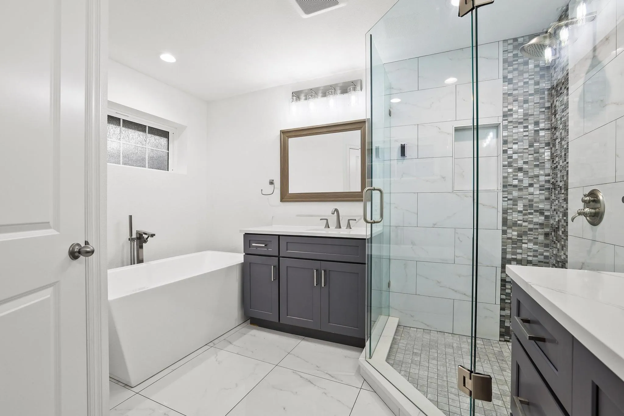 Freestanding soaking tub with floor-mounted fixture, glass-enclosed shower with mosaic tile accent, dual vanities with white countertops, and recessed lighting