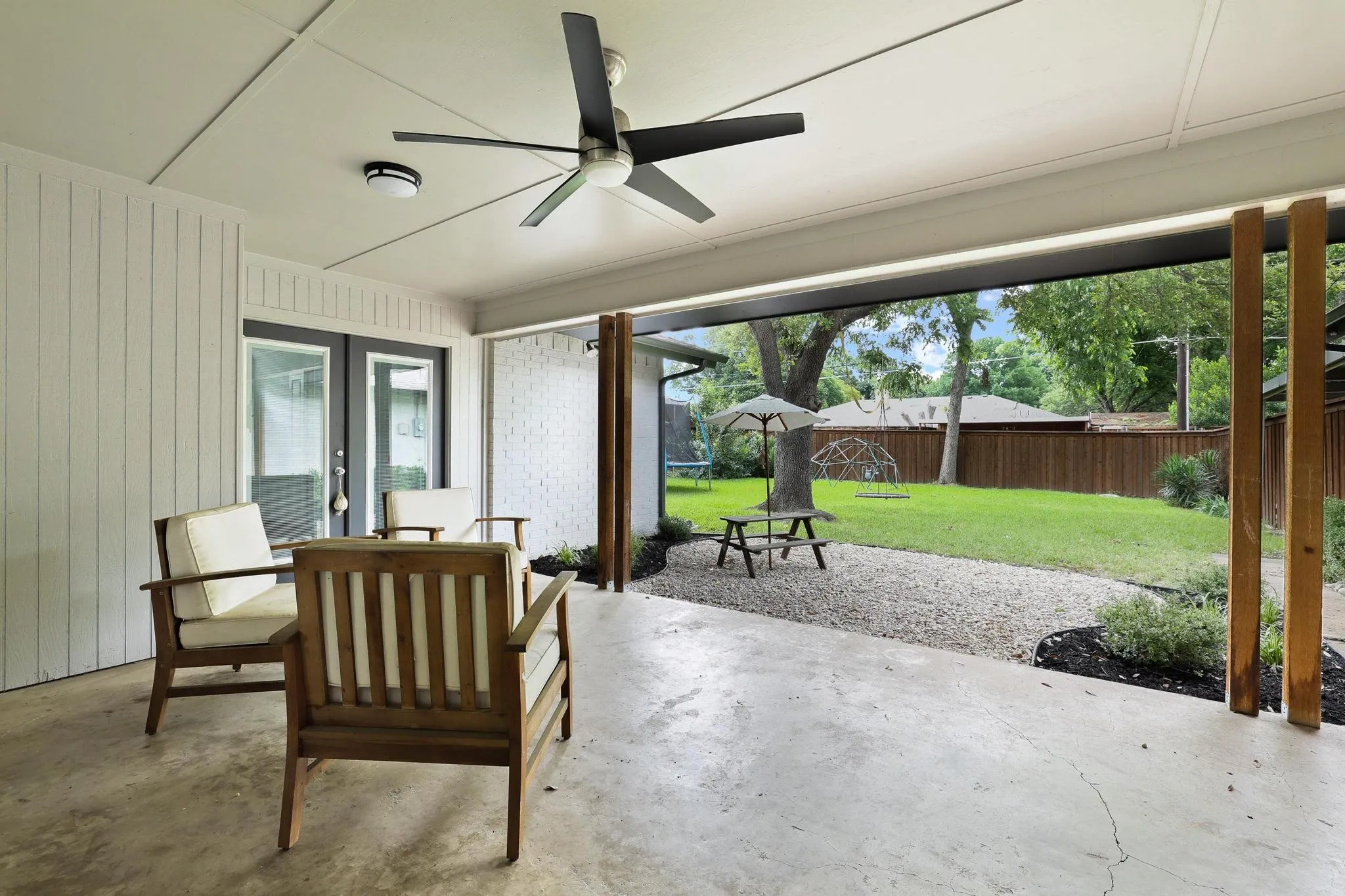 Covered patio with a ceiling fan, concrete flooring, and direct access to a spacious backyard featuring a gravel path and mature trees