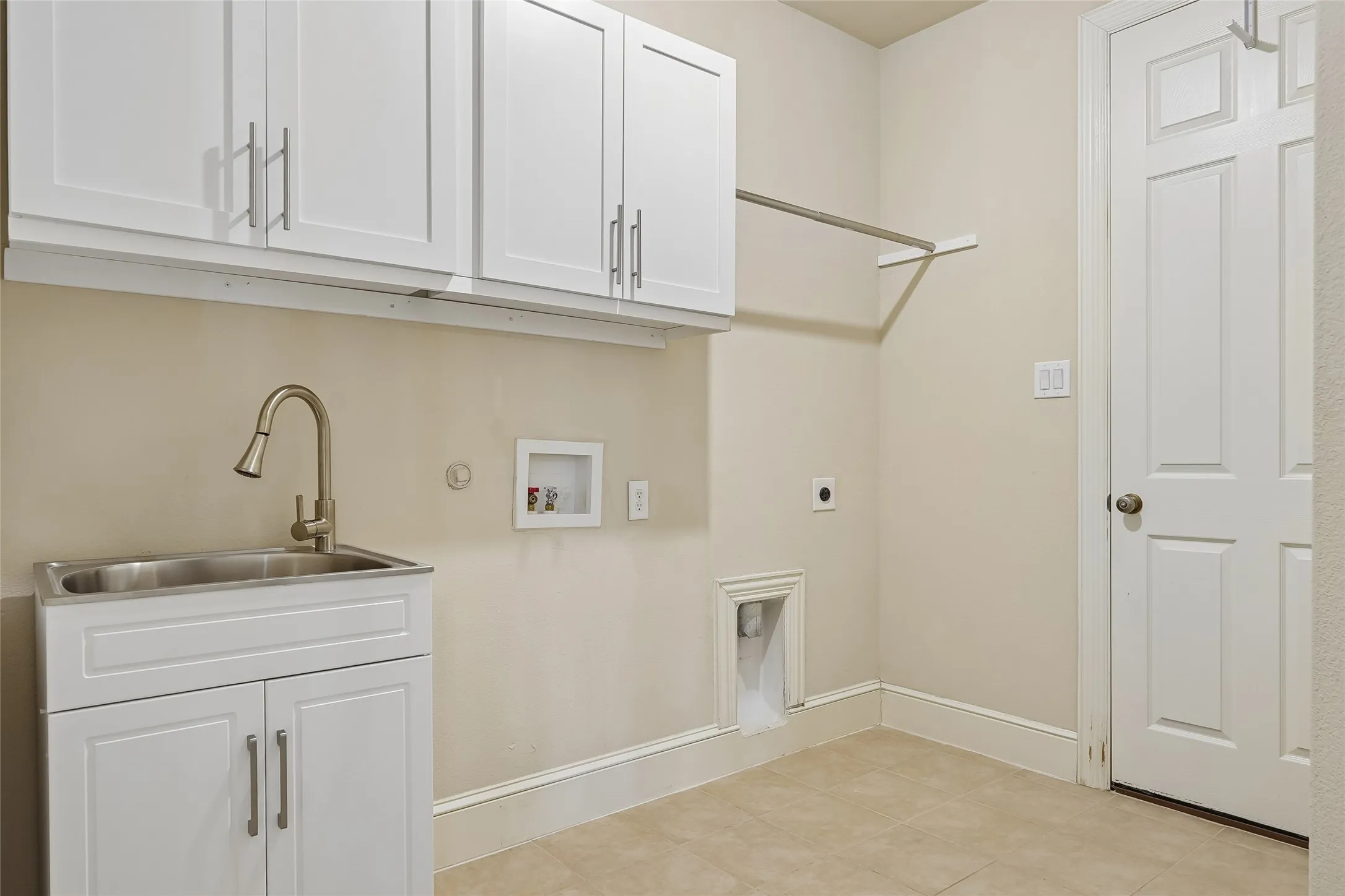 Laundry area featuring overhead white shaker cabinetry, an undermount stainless steel sink with a gooseneck faucet, a built-in hanging rod, and tile flooring