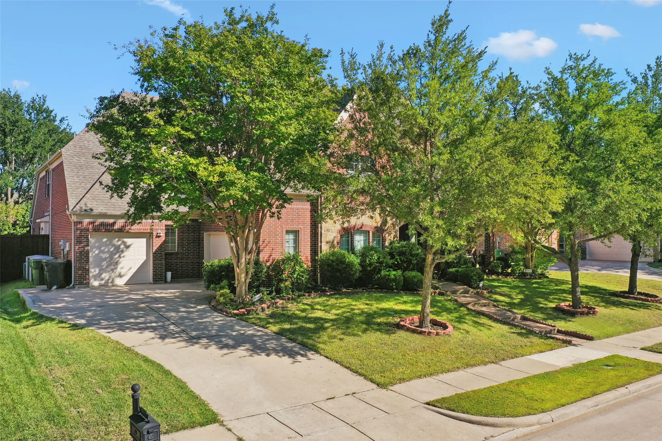 Brick exterior with an attached two-car garage, concrete driveway, front yard landscaping, and mature trees