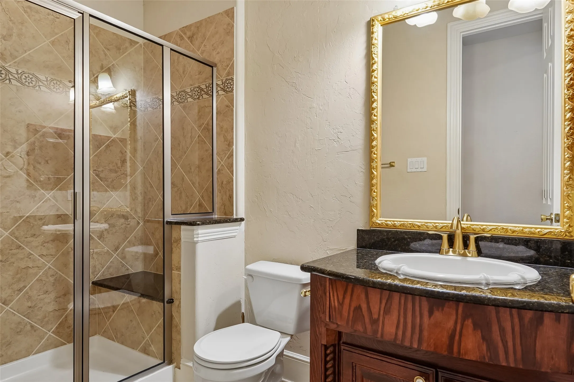 Tiled shower enclosure with glass surround, wood-finish vanity with countertop, decorative mirror, and gold-finish fixtures
