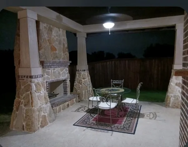 Covered patio at night, with a stone wood-burning fireplace, a ceiling fan, and a fenced backyard