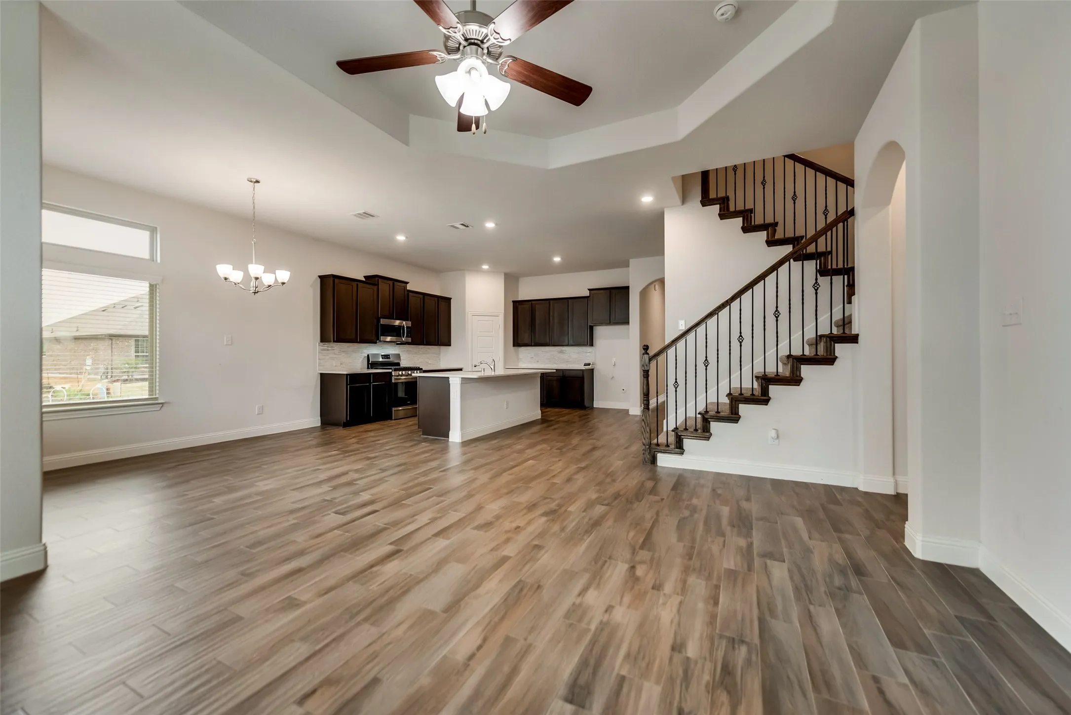Open-concept living space featuring wood-look tile flooring, a kitchen with dark cabinetry and a central island, a staircase with wrought iron balusters, and a tray ceiling with a ceiling fan