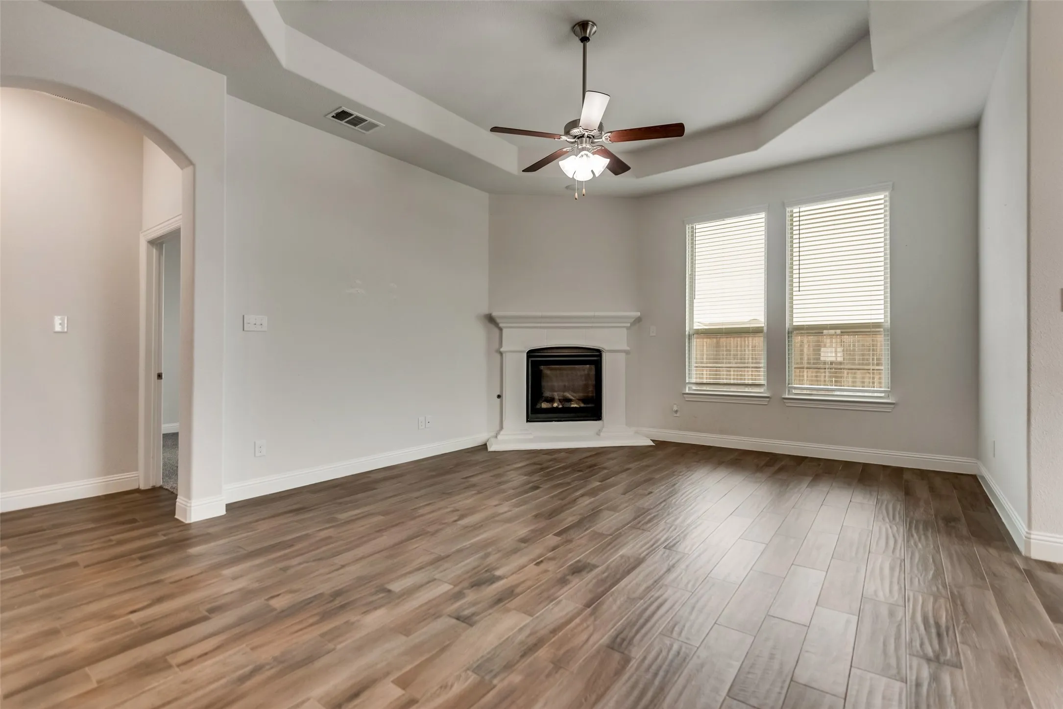 Wood-finish flooring, tray ceiling, ceiling fan, fireplace with mantel, and two windows with blinds