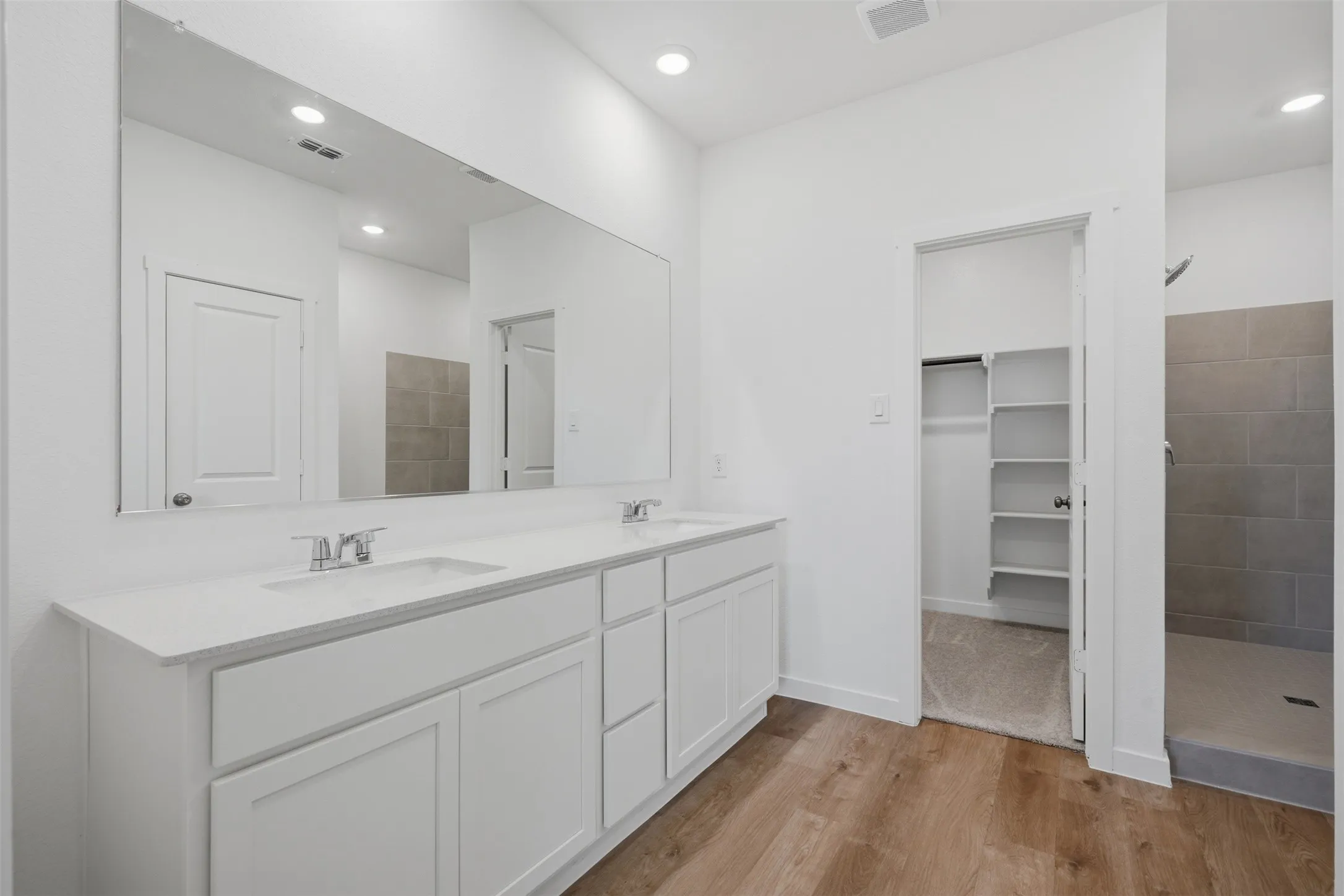 Double vanity with white cabinetry and integrated sinks, wide-plank wood-finish flooring, recessed lighting, and a walk-in shower with large format tile