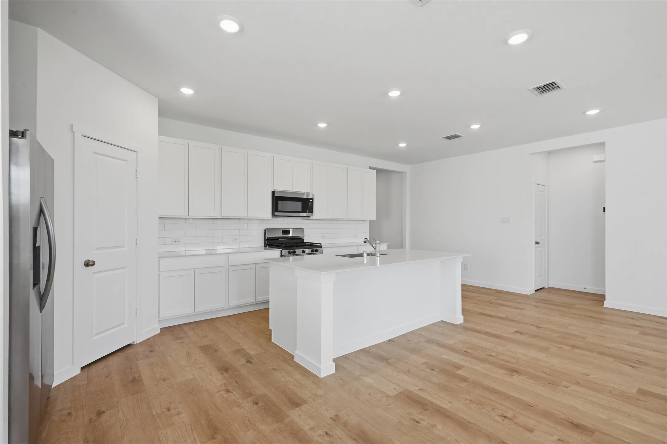 Kitchen featuring an island with a built-in sink, stainless steel appliances, white cabinetry, a subway tile backsplash, and wood-finish flooring
