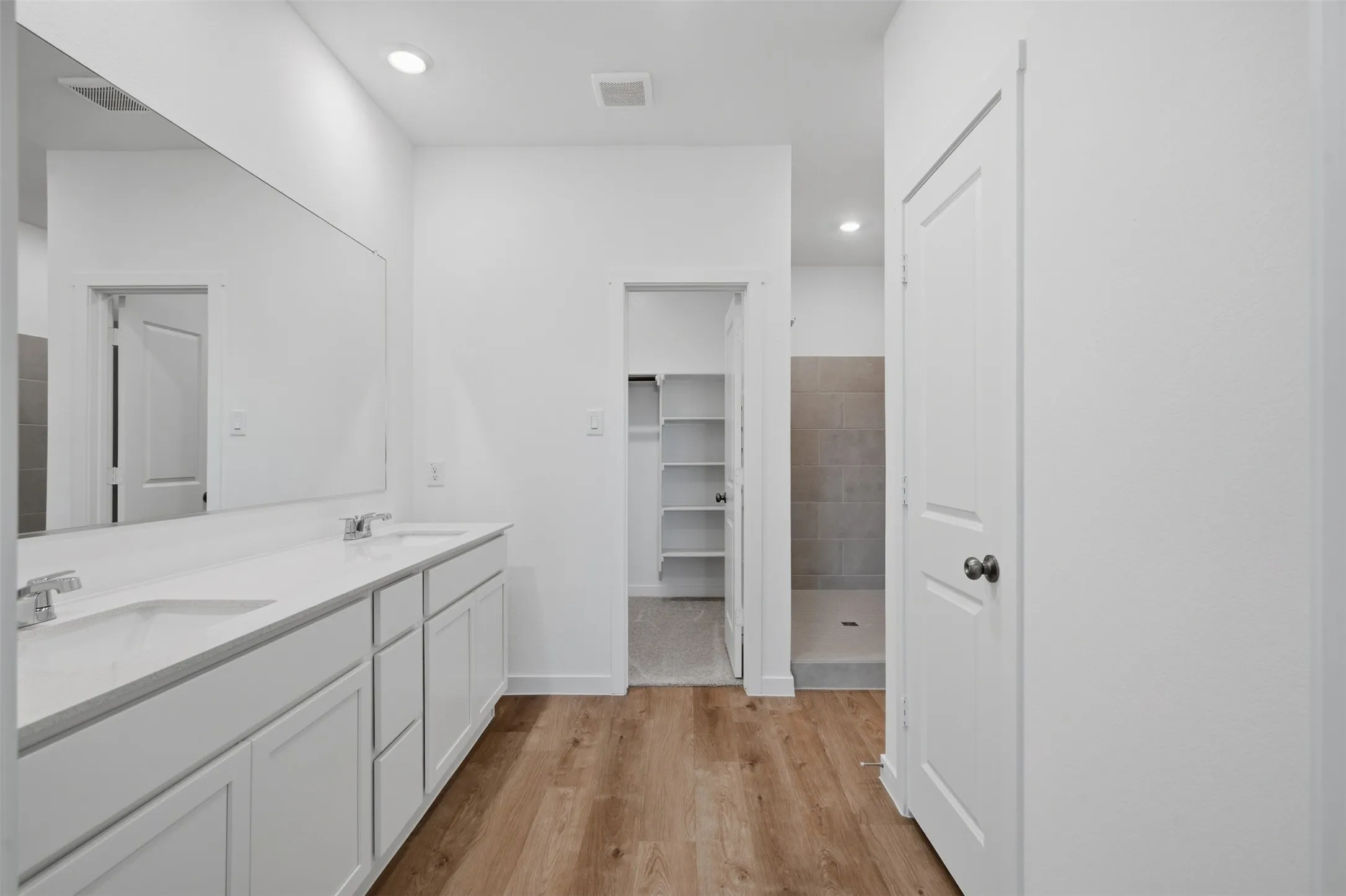 Dual vanity with integrated sinks, wood-finish flooring, recessed lighting, walk-in shower with tiled walls, and a walk-in closet with built-in shelving