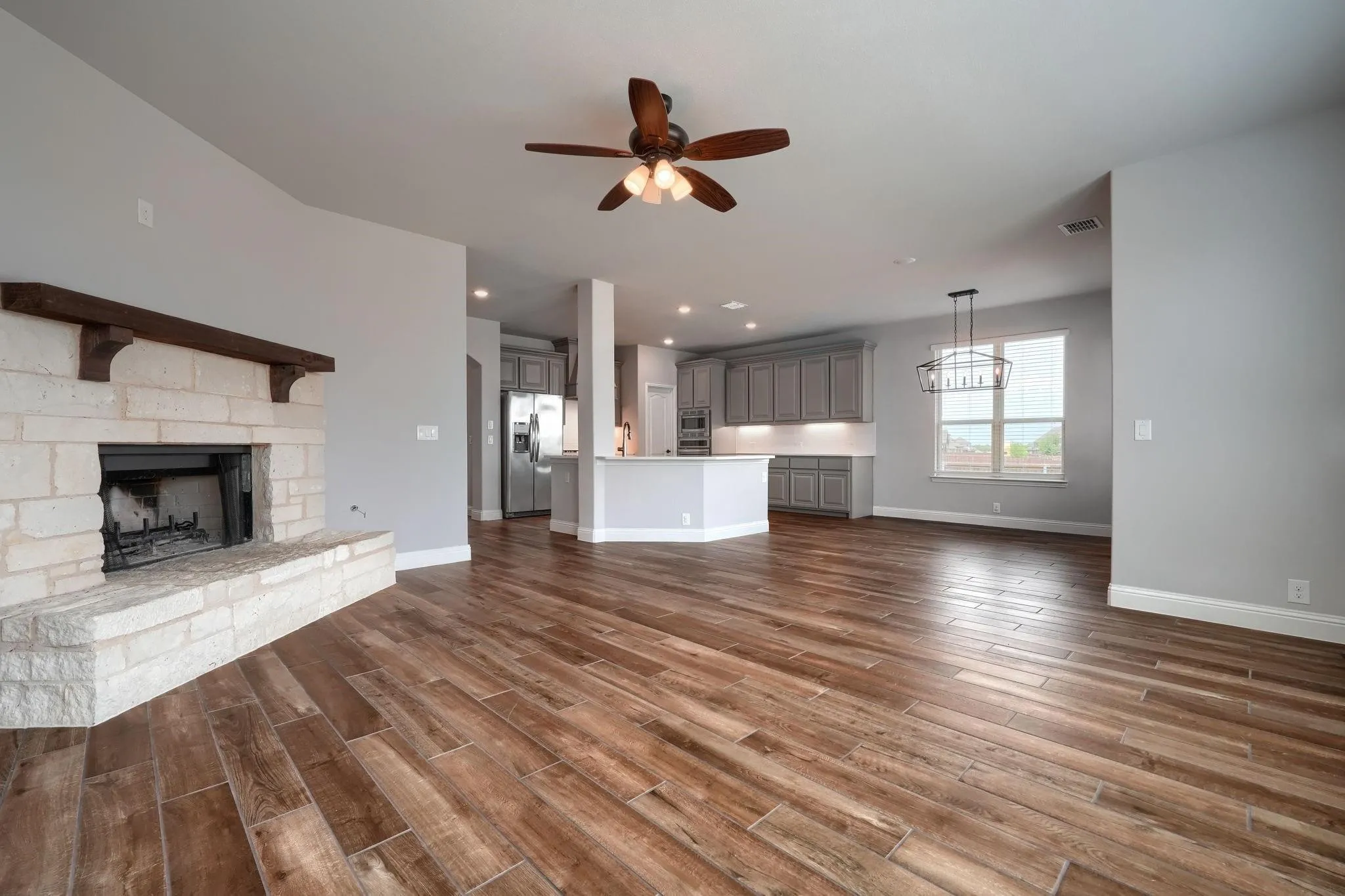 Stone-clad fireplace with a wood mantle, wood-finish flooring, ceiling fan, recessed lighting, and kitchen with gray cabinetry