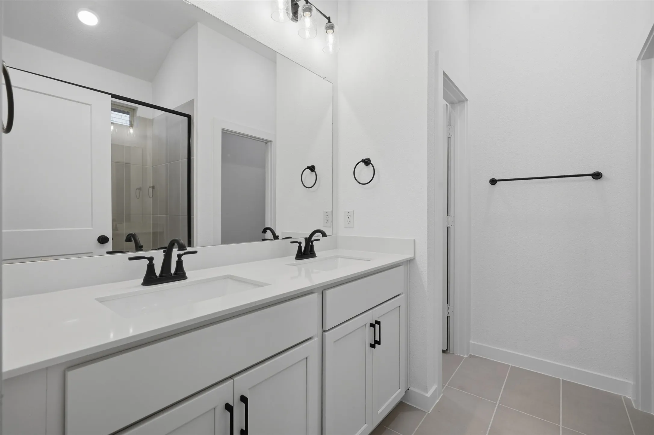 Dual white rectangular undermount sinks set in a light-toned countertop, white shaker-style cabinetry with matte black hardware, large wall-mounted mirror, and a clear glass shower enclosure with black framing