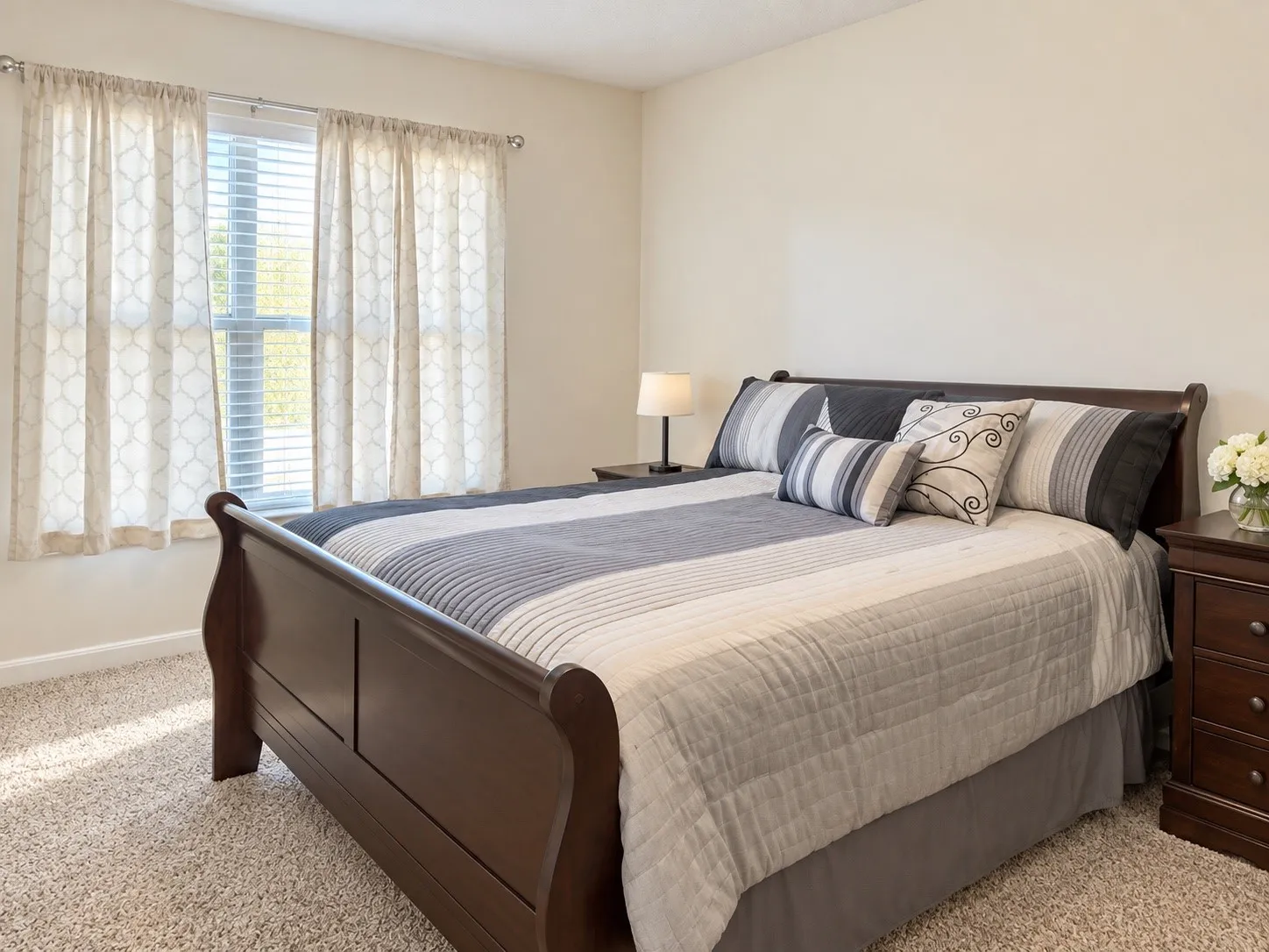 Window with blinds, warm neutral wall color, plush carpet flooring, dark wood sleigh bed frame, dark wood nightstand with drawers