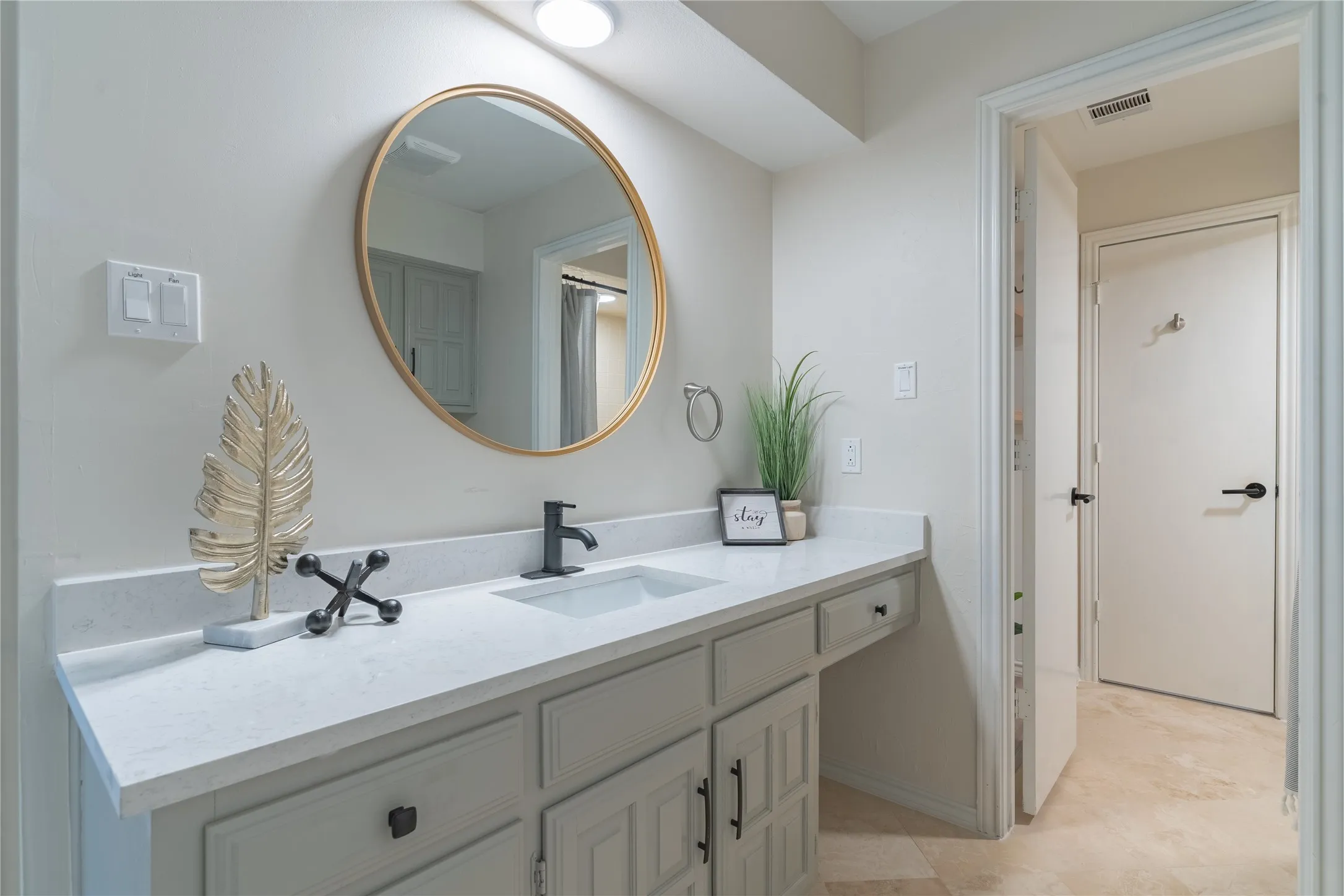 Vanity with a gold-framed circular mirror, white countertop, matte black faucet, and light-colored cabinetry