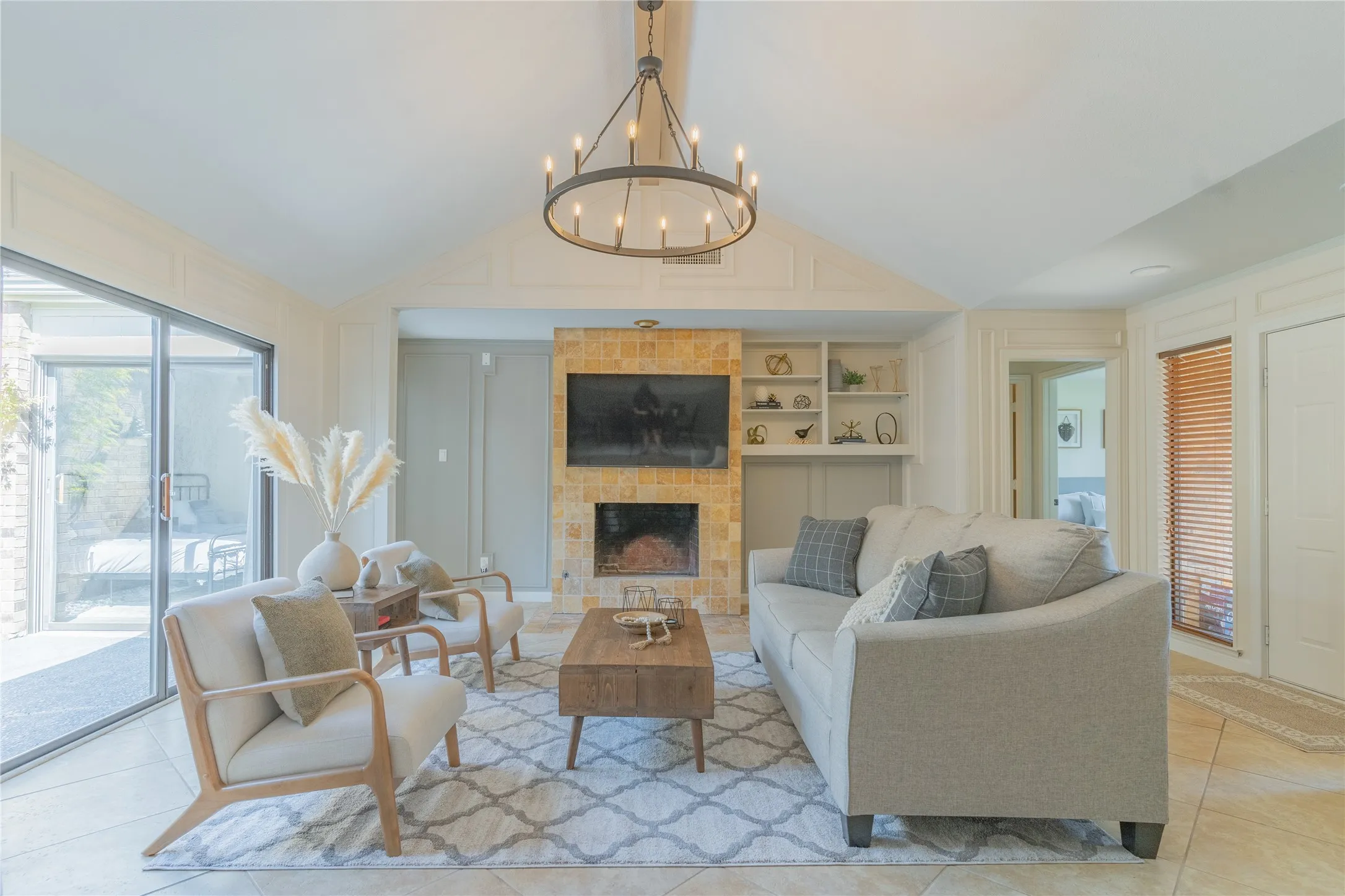 Vaulted ceiling with an iron chandelier, stacked stone fireplace, built-in shelving, tile flooring, and sliding glass doors
