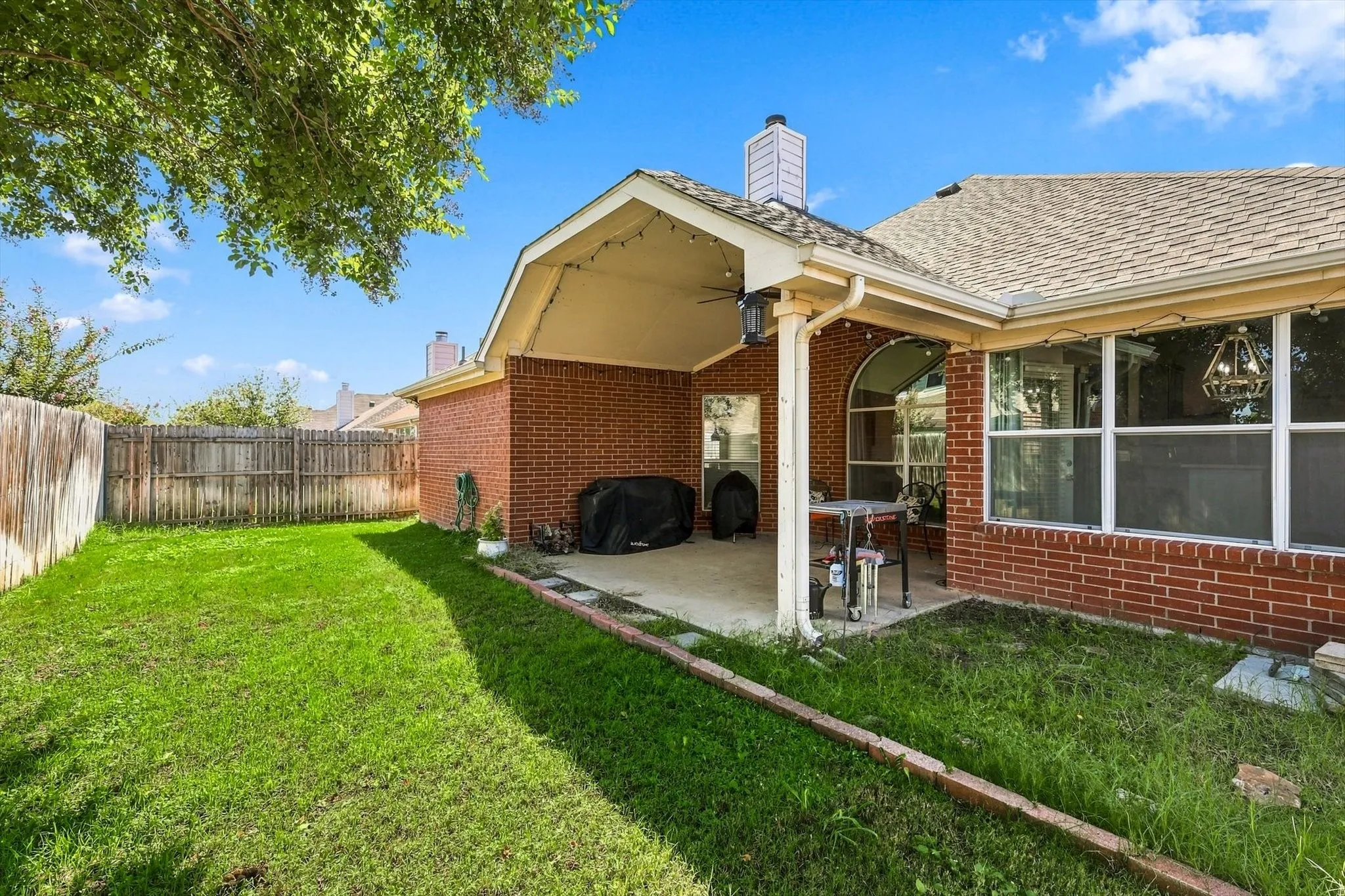 Expansive backyard featuring a covered patio, brick exterior, fencing, and a shingled roof