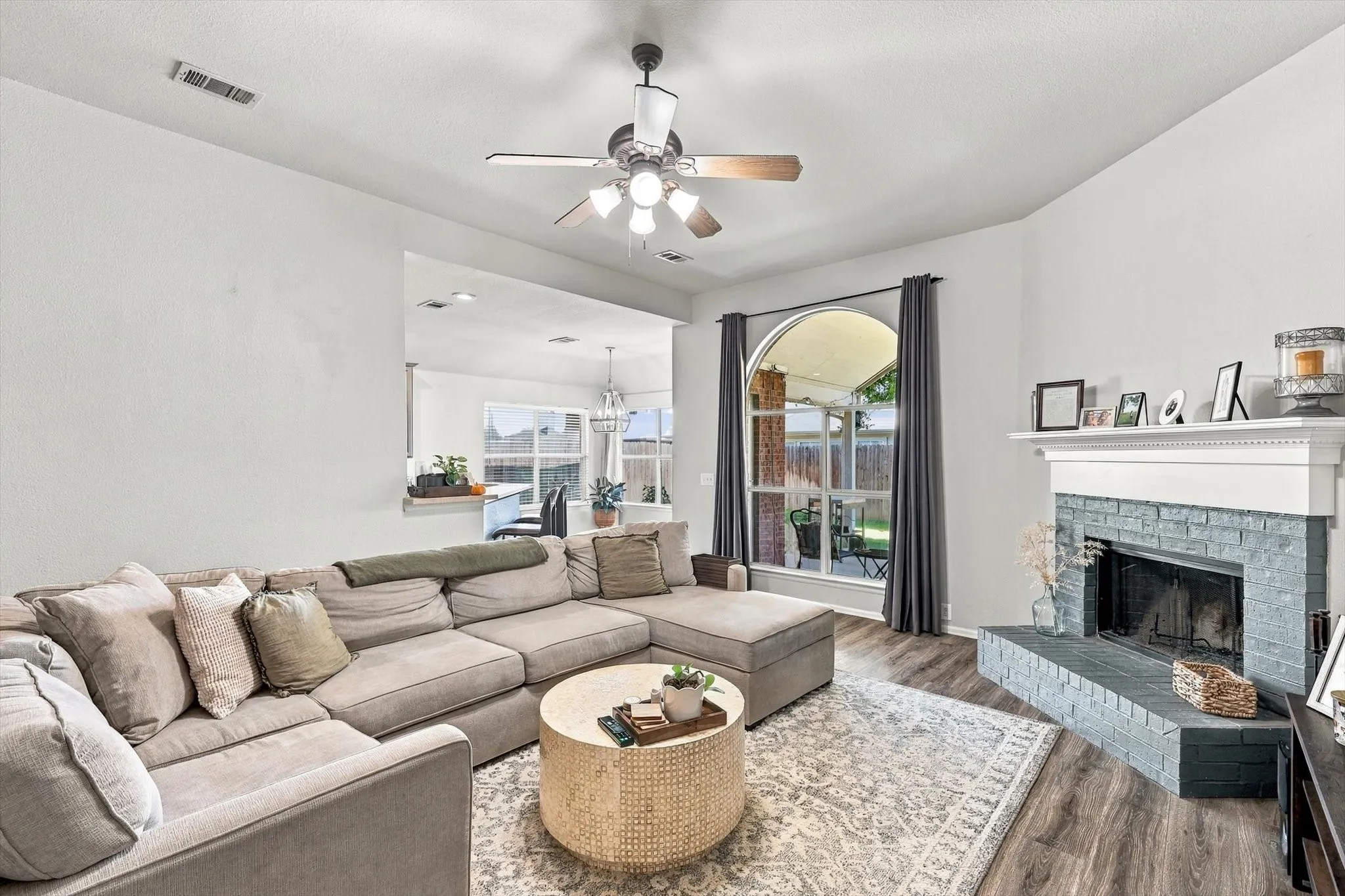 Living area featuring wood-finish flooring, a ceiling fan, and a fireplace with a painted brick surround and white mantel