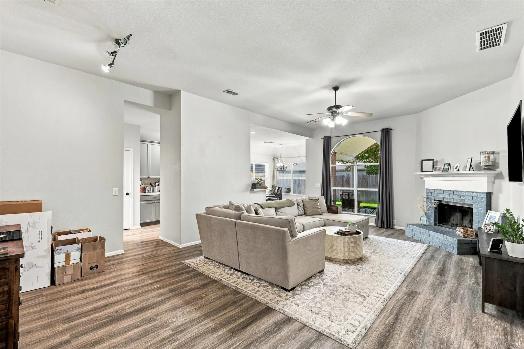 Wood-finish flooring throughout the living space, a fireplace with a light-colored brick surround, a large arched window, and a ceiling fan with integrated lighting