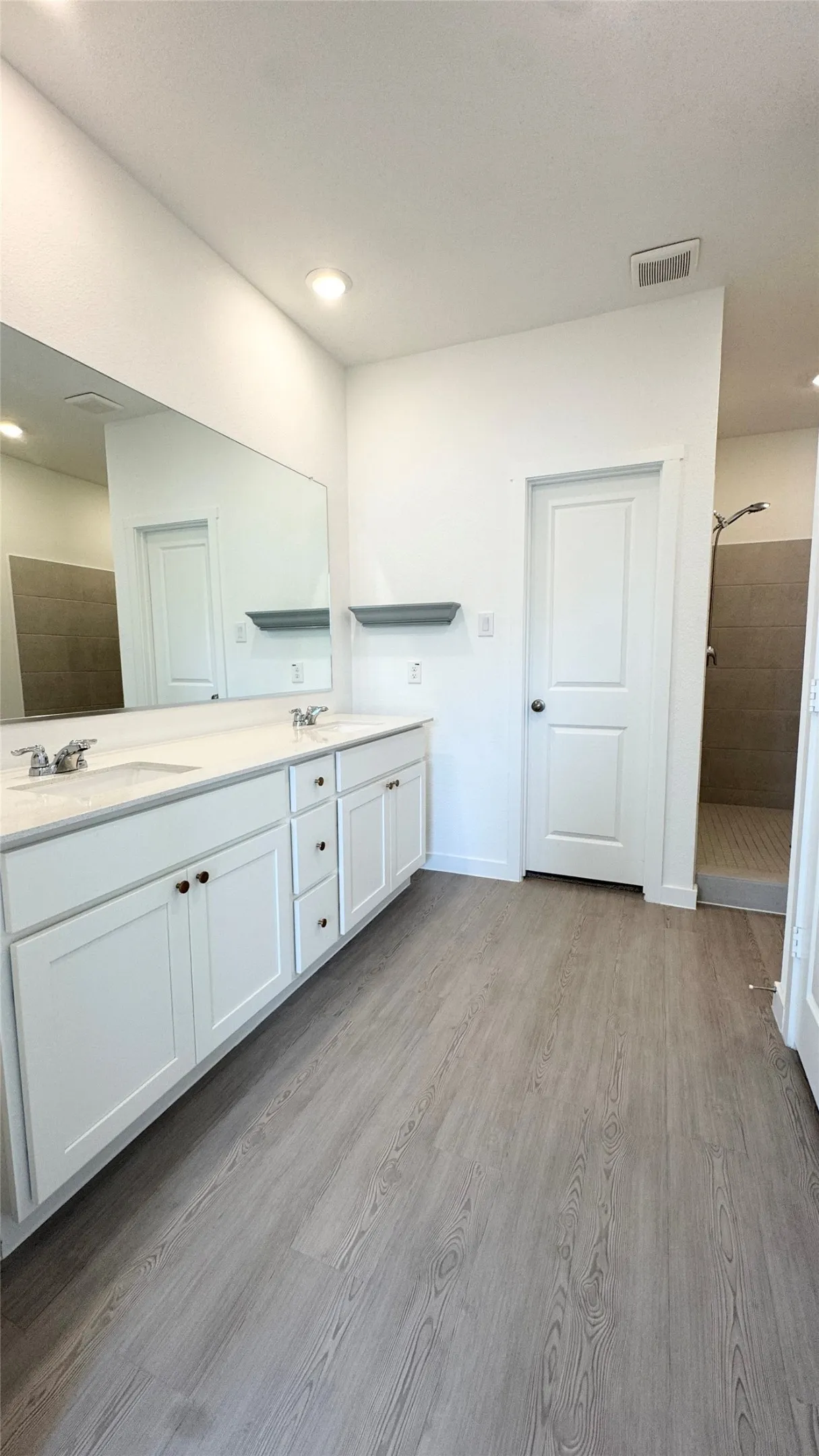 Dual vanity with white cabinetry and integrated sinks, a large wall-mounted mirror, wood-finish flooring, and a walk-in shower with tiled walls