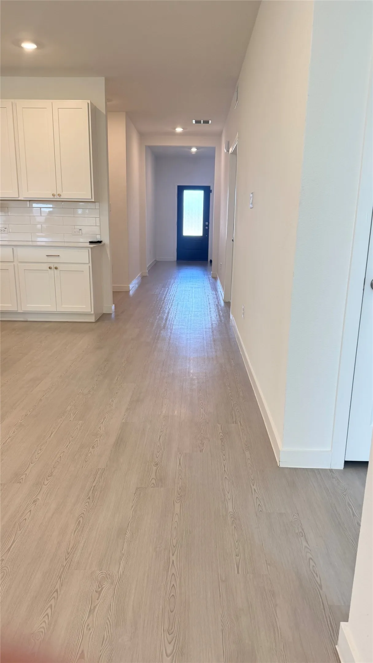 Wood-finish flooring throughout, white cabinetry with silver hardware, subway tile backsplash, recessed lighting, and a dark entry door with frosted glass
