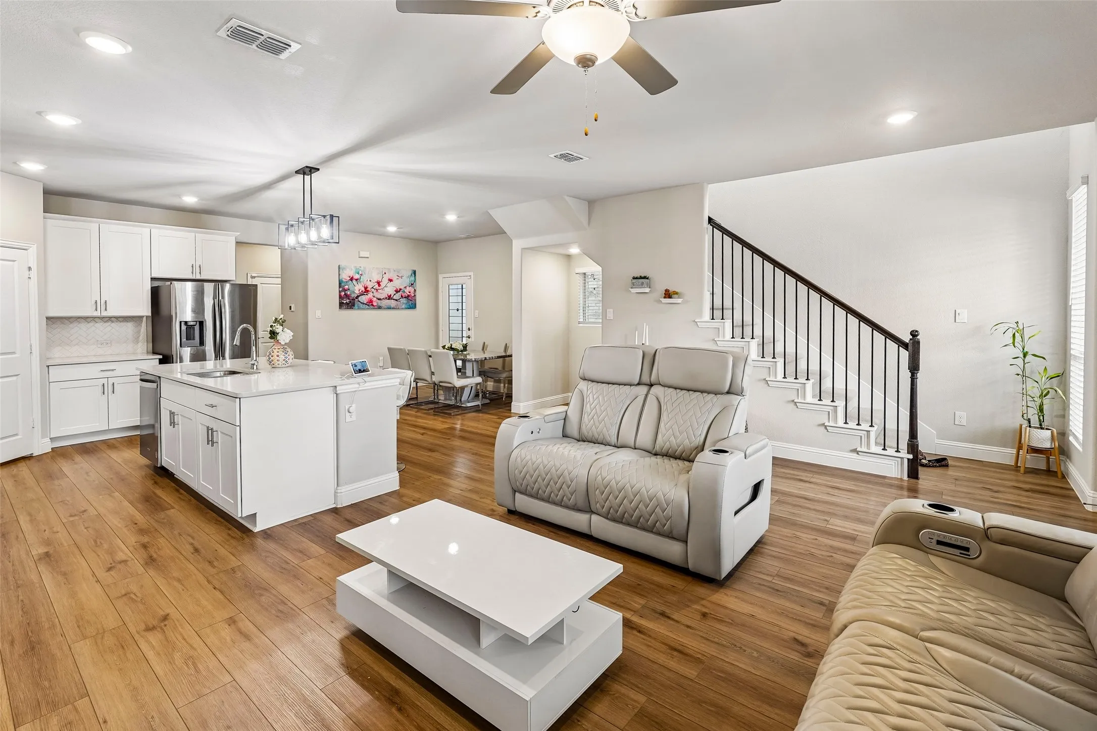 Open-concept living space featuring wood-finish flooring, a kitchen island with a sink, white cabinetry, stainless steel appliances, and a staircase with wrought iron balusters