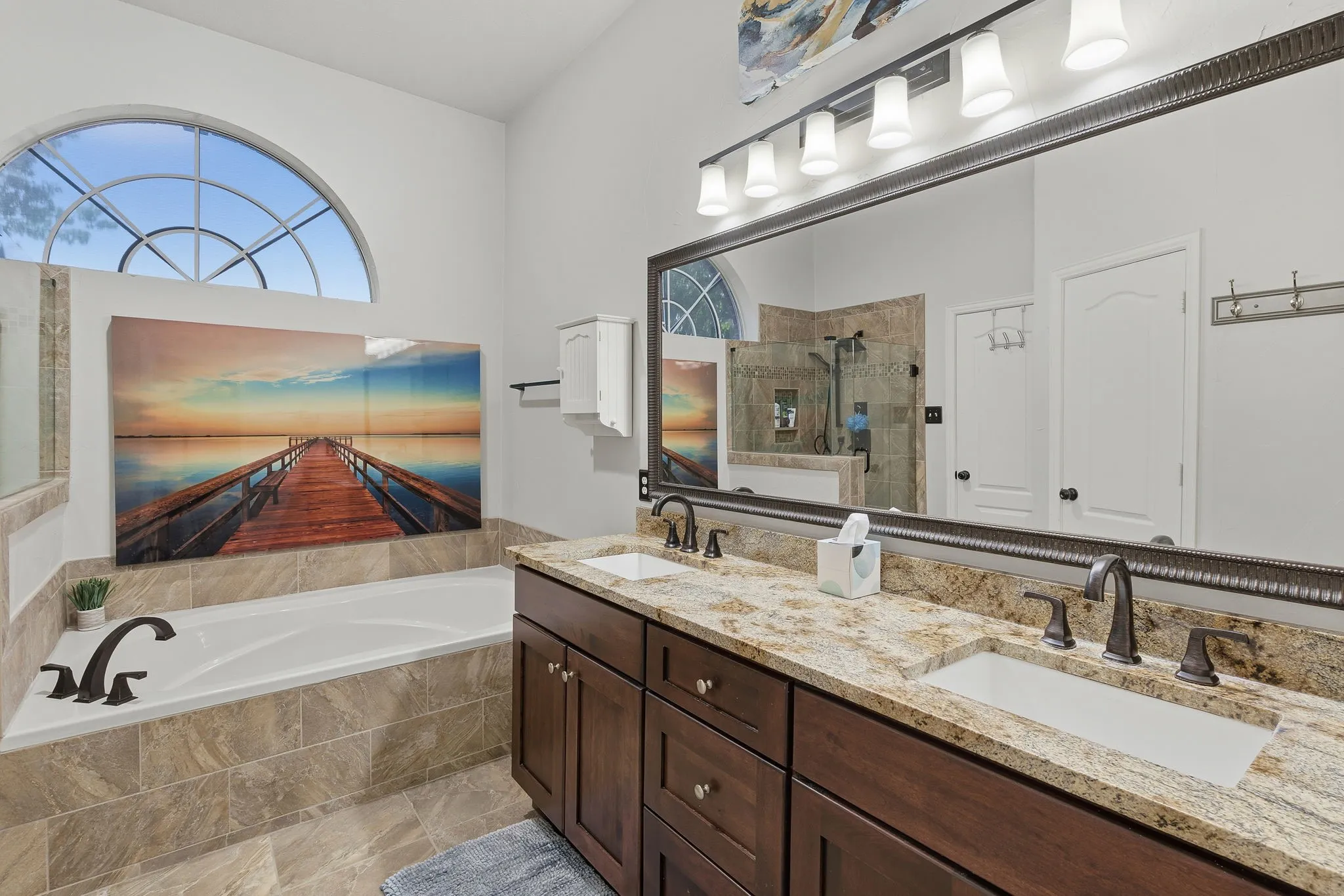 Dual vanity with granite countertops and undermount sinks, large framed mirror with integrated lighting, built-in soaking tub with tiled surround, and arched window