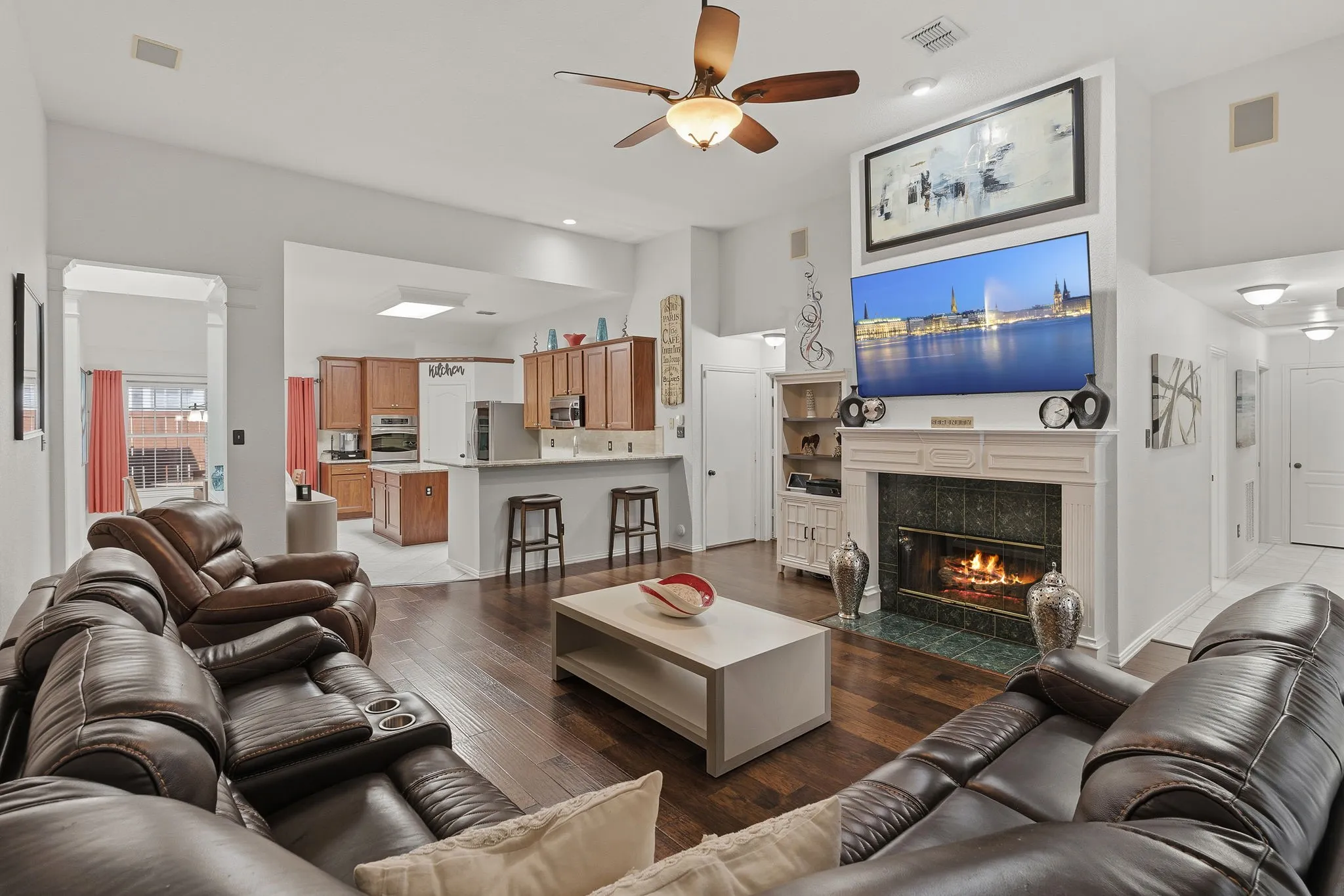 Vaulted ceiling with a ceiling fan, fireplace with a decorative mantel and tile surround, wood-finish flooring, recessed lighting, and a kitchen with wood cabinetry and a breakfast bar