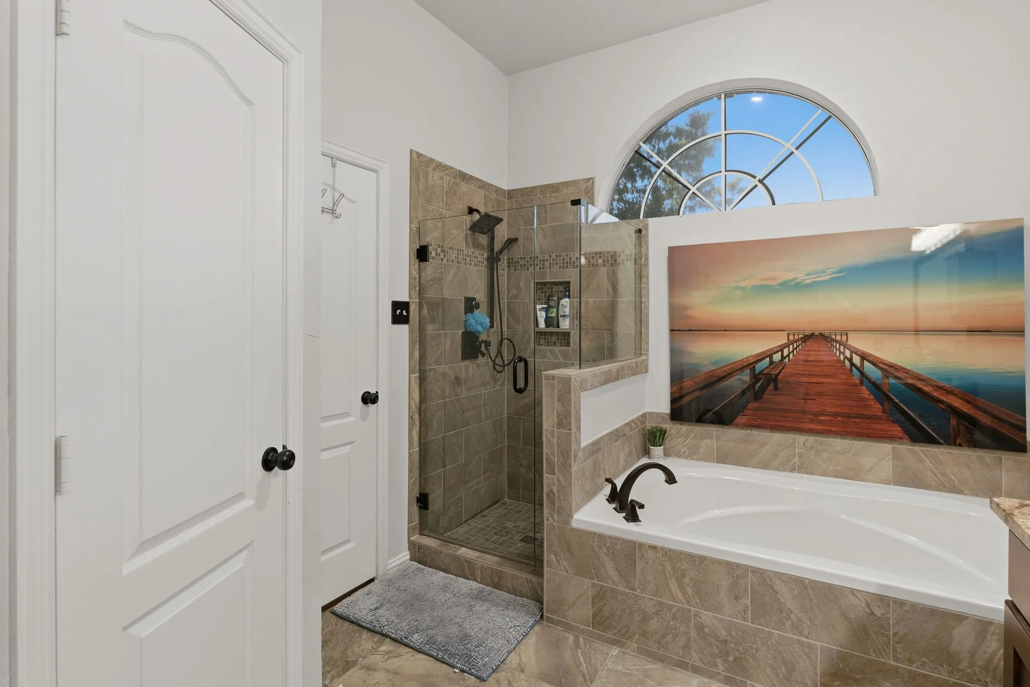 Tile-surround soaking tub with bronze-finish fixtures, a tiled shower enclosure with a glass door, and a decorative arched window