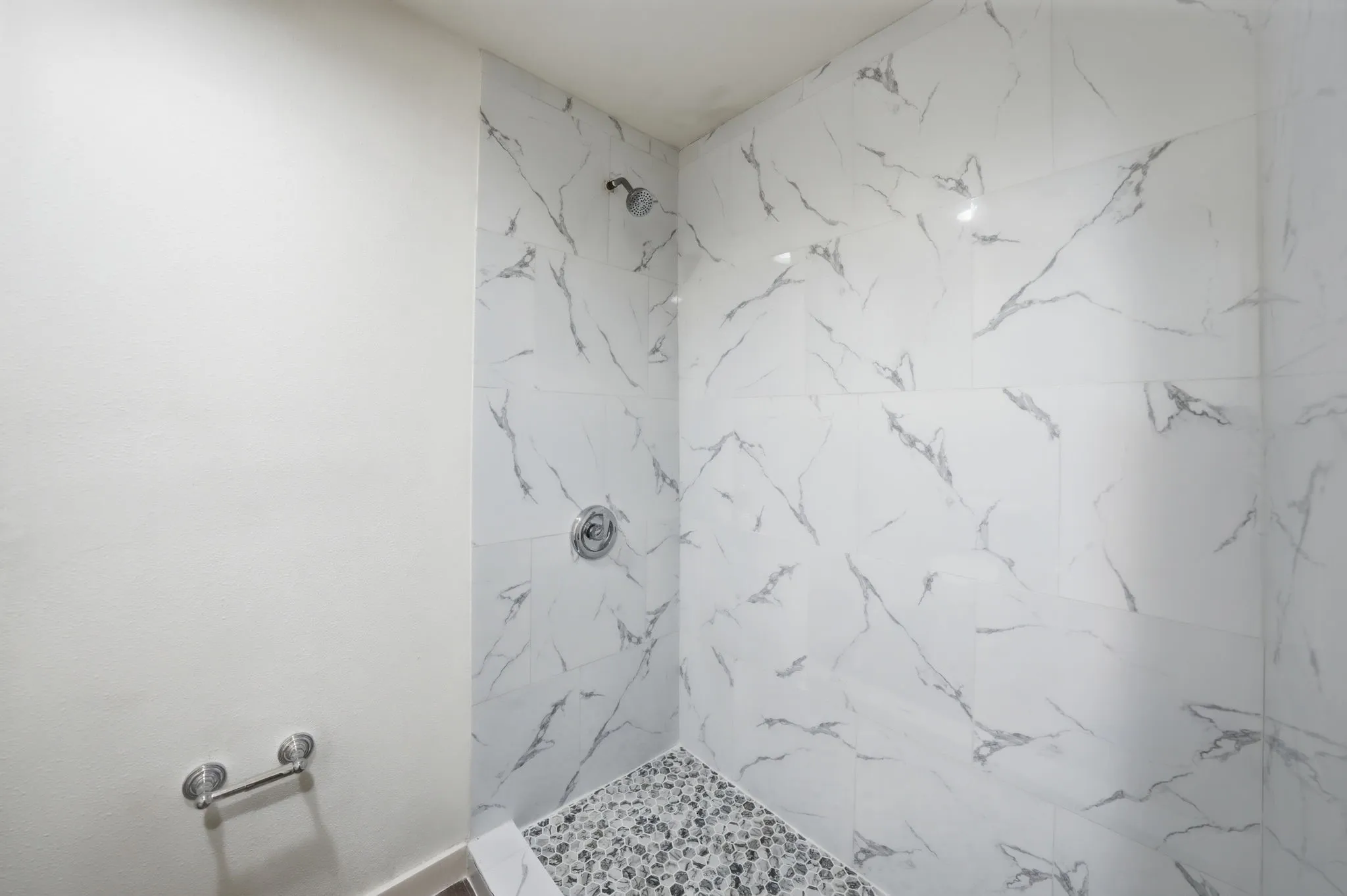 Shower enclosure featuring large format marble-look tile walls, a pebble-stone floor, and a polished chrome shower head and controls