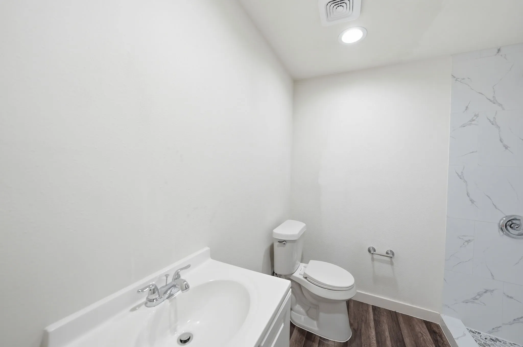 Bathroom featuring a white vanity with an integrated sink, wood-finish flooring, a white toilet, and a shower with white marble-patterned tile