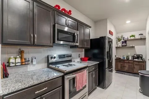 Dark wood-finish cabinetry, stainless steel microwave, stainless steel range, light-toned tile backsplash, and light-toned tile flooring