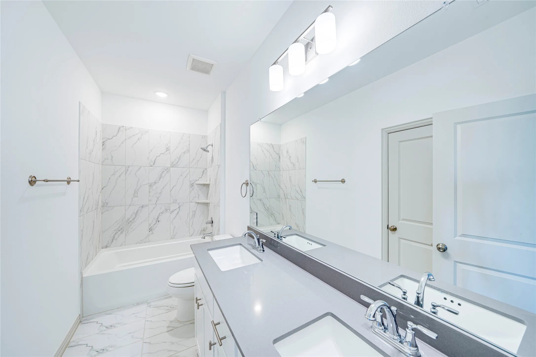 Dual vanity with undermount sinks and chrome fixtures, large wall-mounted mirror, three-light vanity bar, integrated tub/shower with tiled surround, and white floor tiling