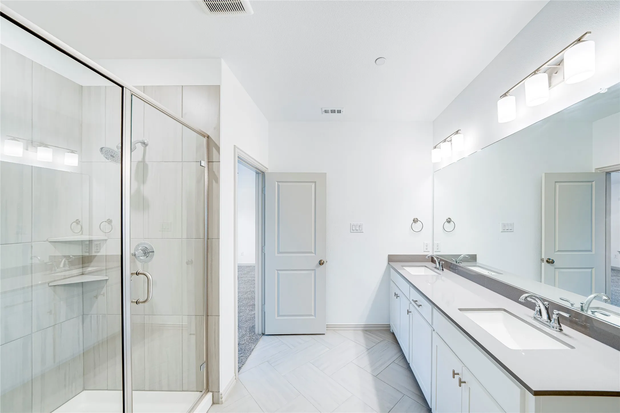 Glass-enclosed shower with tiled walls and built-in shelving, dual vanity with undermount sinks and extensive countertop space, full-width mirror, and tiled flooring