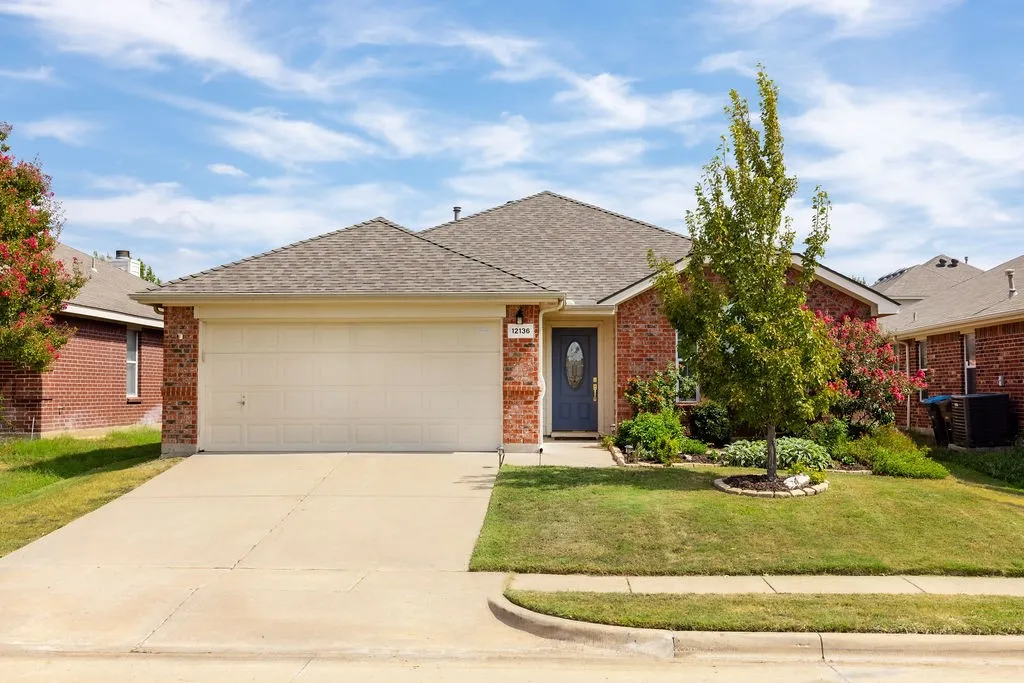 Brick exterior with a gray shingle roof, attached garage, concrete driveway, and established front landscaping
