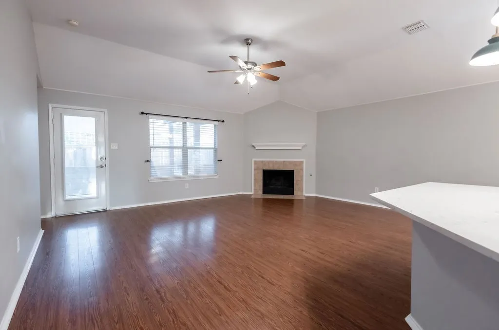 Vaulted ceiling with a ceiling fan, wood-finish flooring, fireplace with a tiled surround and mantel, and an exterior door with a full-light panel