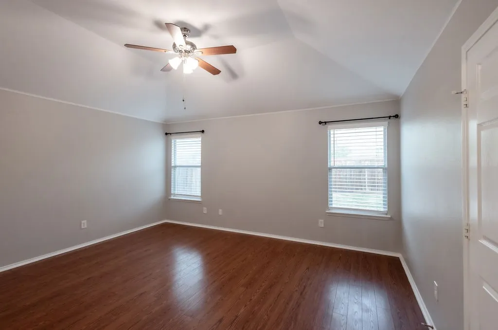 Vaulted ceiling, wood-finish flooring, two windows with blinds, ceiling fan with integrated lighting, and a white paneled door