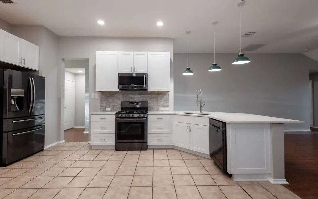 White shaker cabinetry, black stainless steel appliances, light-toned tile flooring, and an island with a sink and pendant lighting