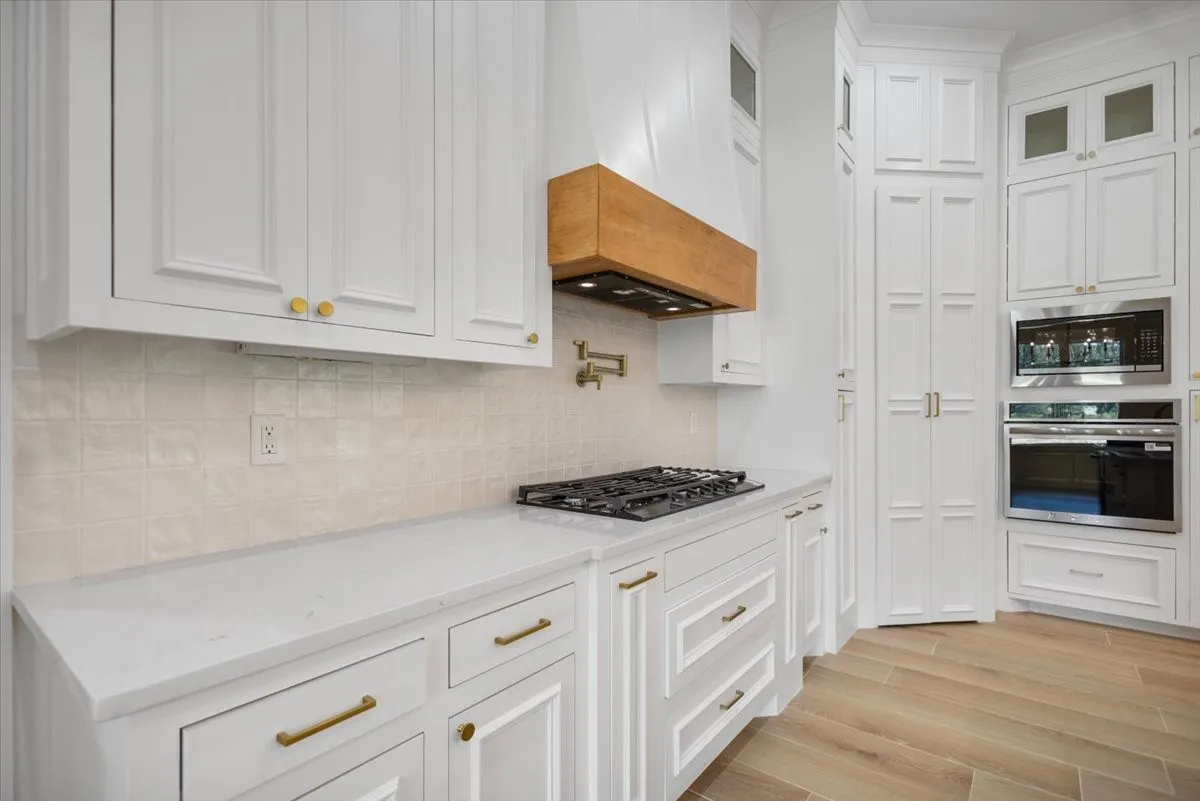 White cabinetry with gold-finish hardware, light-toned backsplash, gas cooktop, built-in oven, and wood-finish flooring