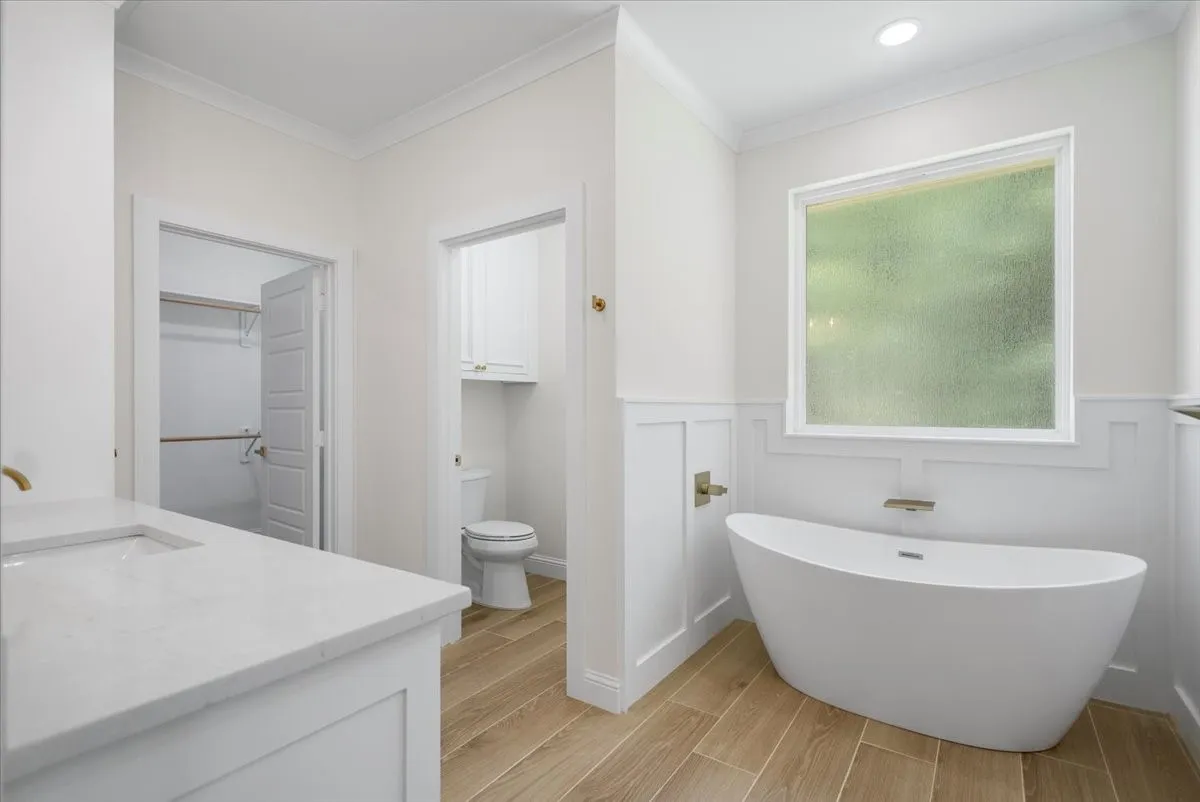Freestanding bathtub with wall-mounted faucet, wood-finish flooring, white paneling, stone countertop, and recessed lighting