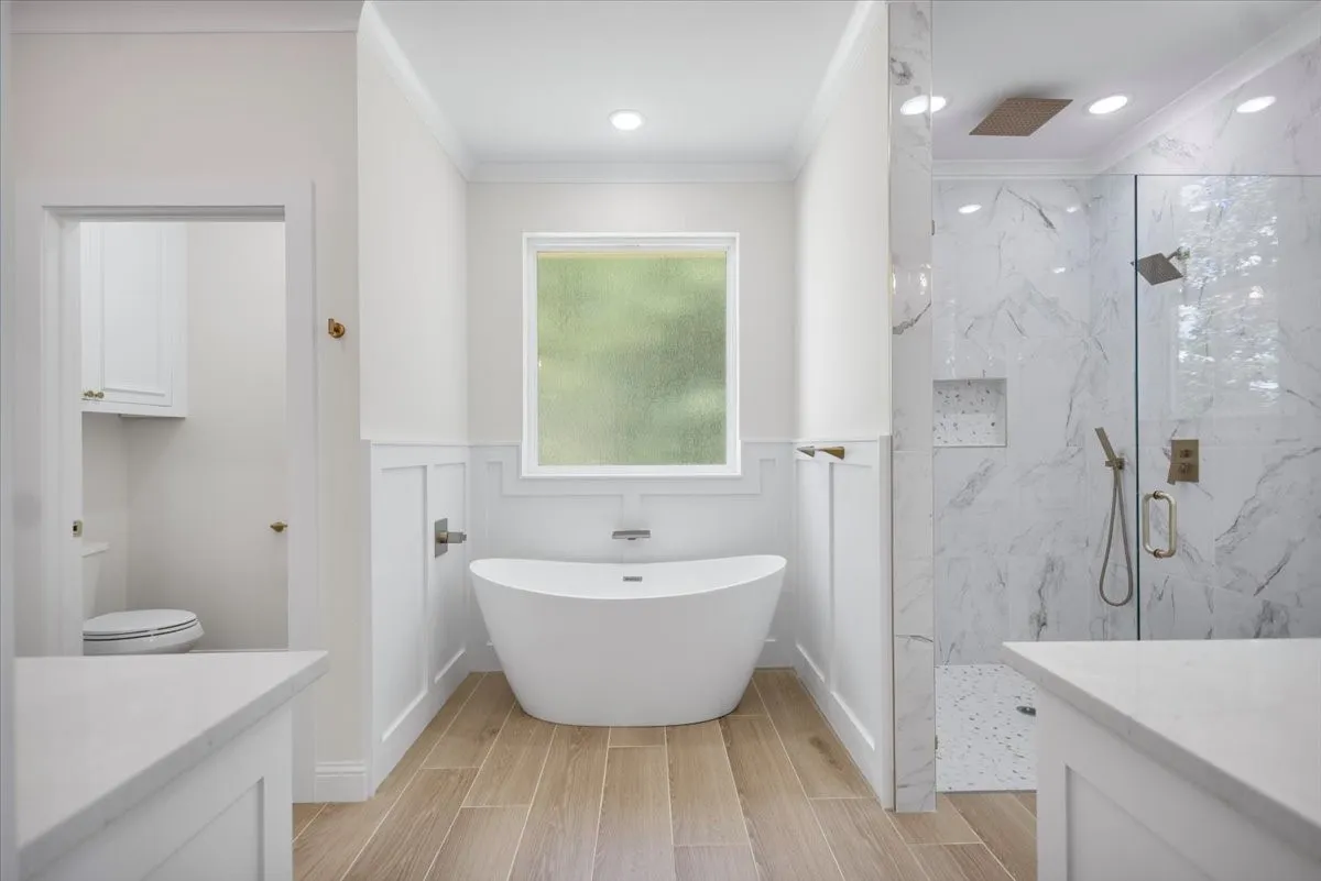 Freestanding soaking tub, wood-finish flooring, glass-enclosed shower with tiled walls, dual vanity countertops, and wainscoting