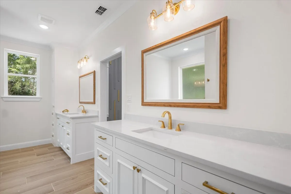 Dual vanity with white cabinetry and gold-toned hardware, white solid surface countertops, gold-toned wall-mounted lighting, and wood-finish flooring