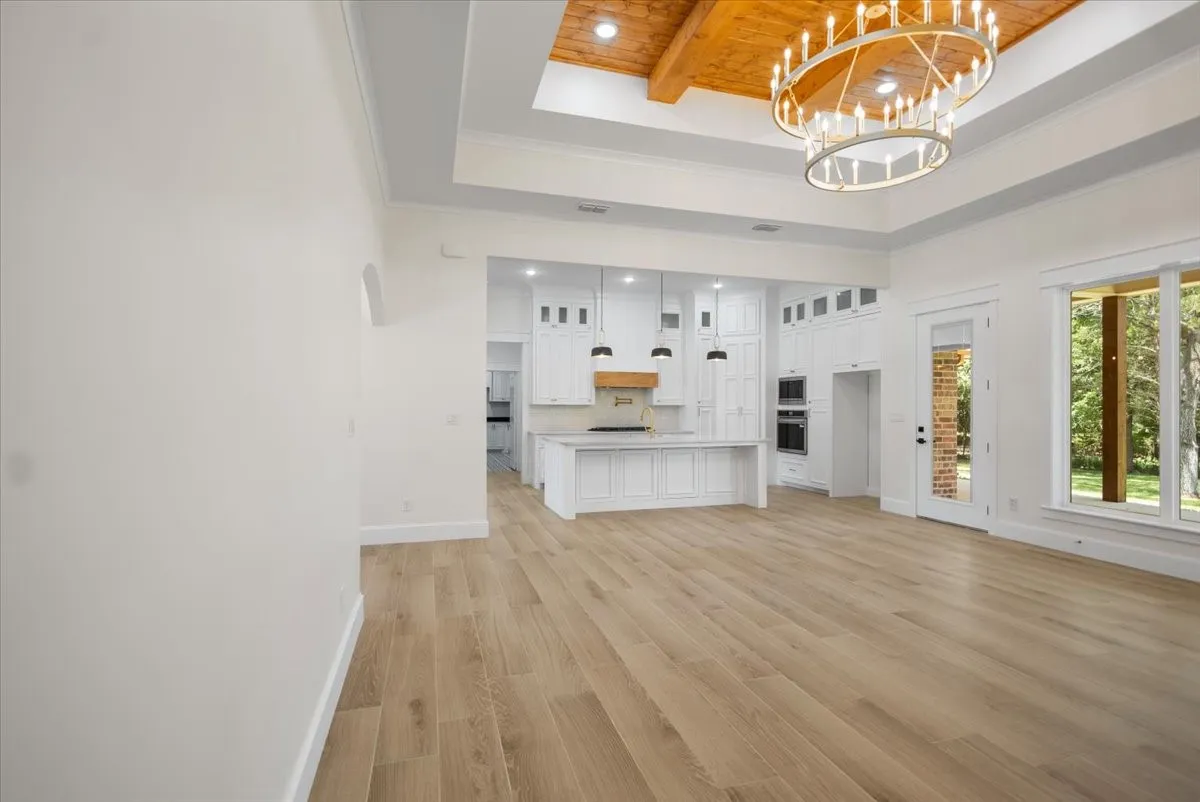 Wood-finish flooring throughout, tray ceiling with wood-paneled accent, large chandelier, white kitchen cabinetry with island, and large windows