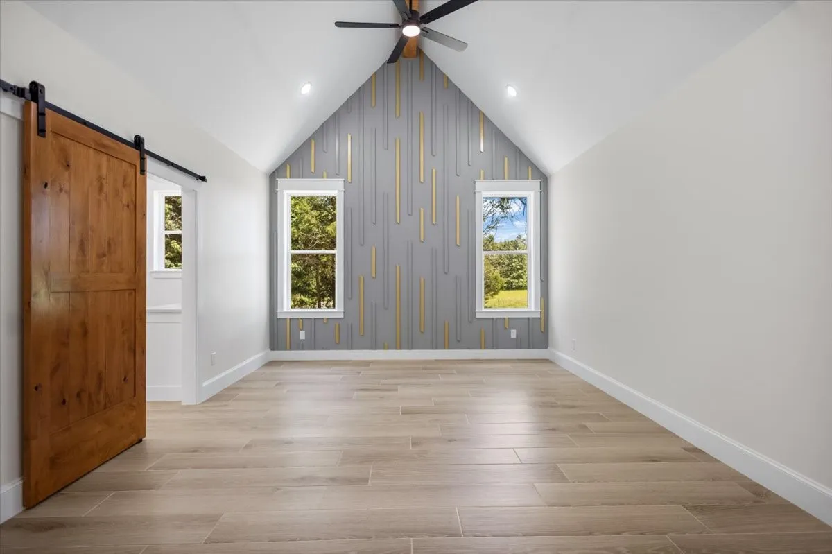 Vaulted ceiling featuring a ceiling fan, wood-finish flooring, accent wall with vertical paneling, and a wood barn door