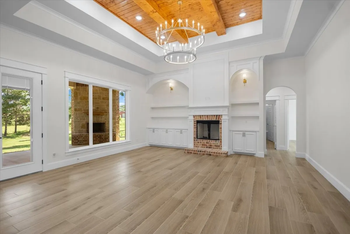Wood-finish flooring, a brick fireplace with built-in shelving, tray ceiling with wood paneling, a large chandelier, and expansive windows